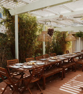 An outdoor dining area with a long wooden table set with plates, utensils, and glasses under a covered patio, surrounded by greenery at Grace Troutman's educational photography retreat in California.