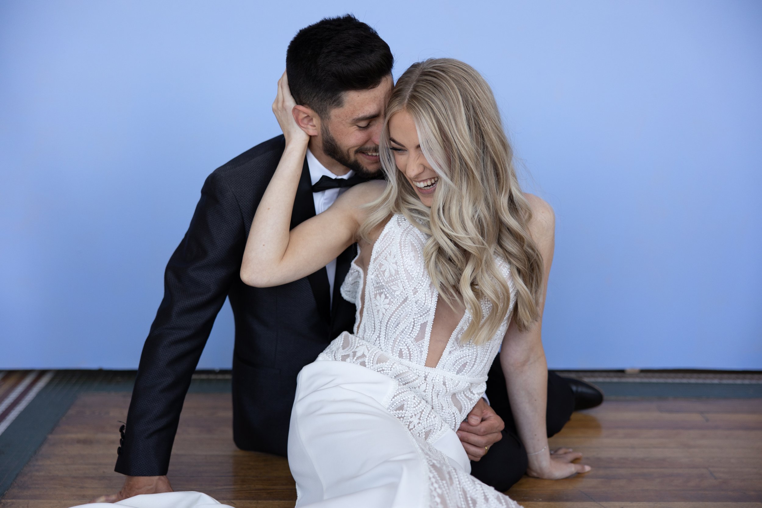 A couple dressed in wedding attire, a man in a black tuxedo and a woman in a white wedding dress, sharing a joyful moment on the floor.