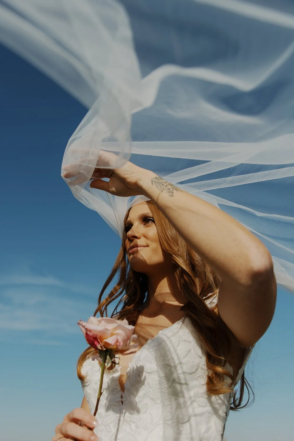 A bride with long red hair holds a pink rose and shields her eyes with a sheer white veil against a bright blue sky.