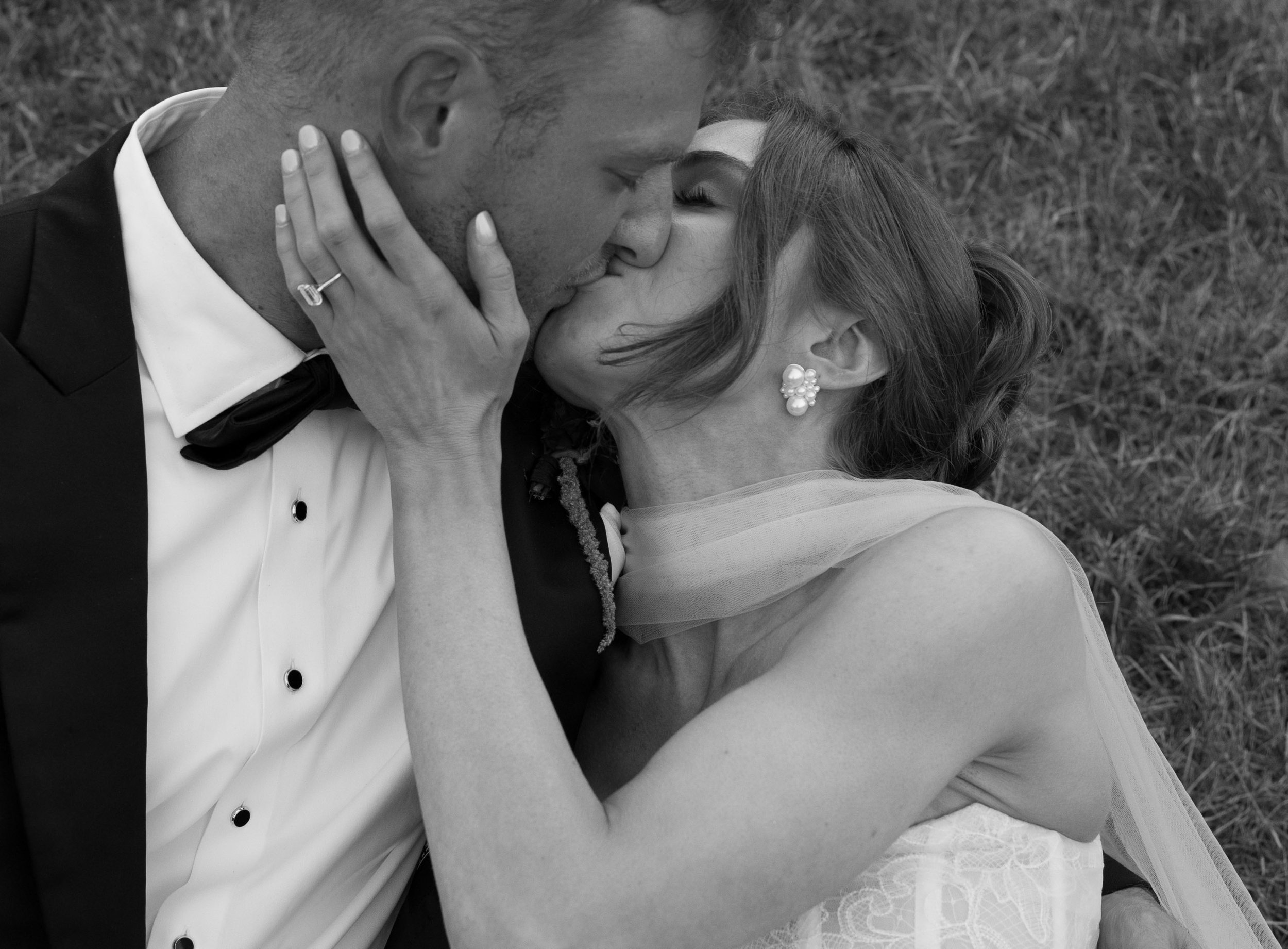A black-and-white photo of a bride and groom sharing a kiss outdoors, with grass in the background. The groom is wearing a tuxedo and the bride has her hand on his face, showing her wedding ring and pearl earrings.