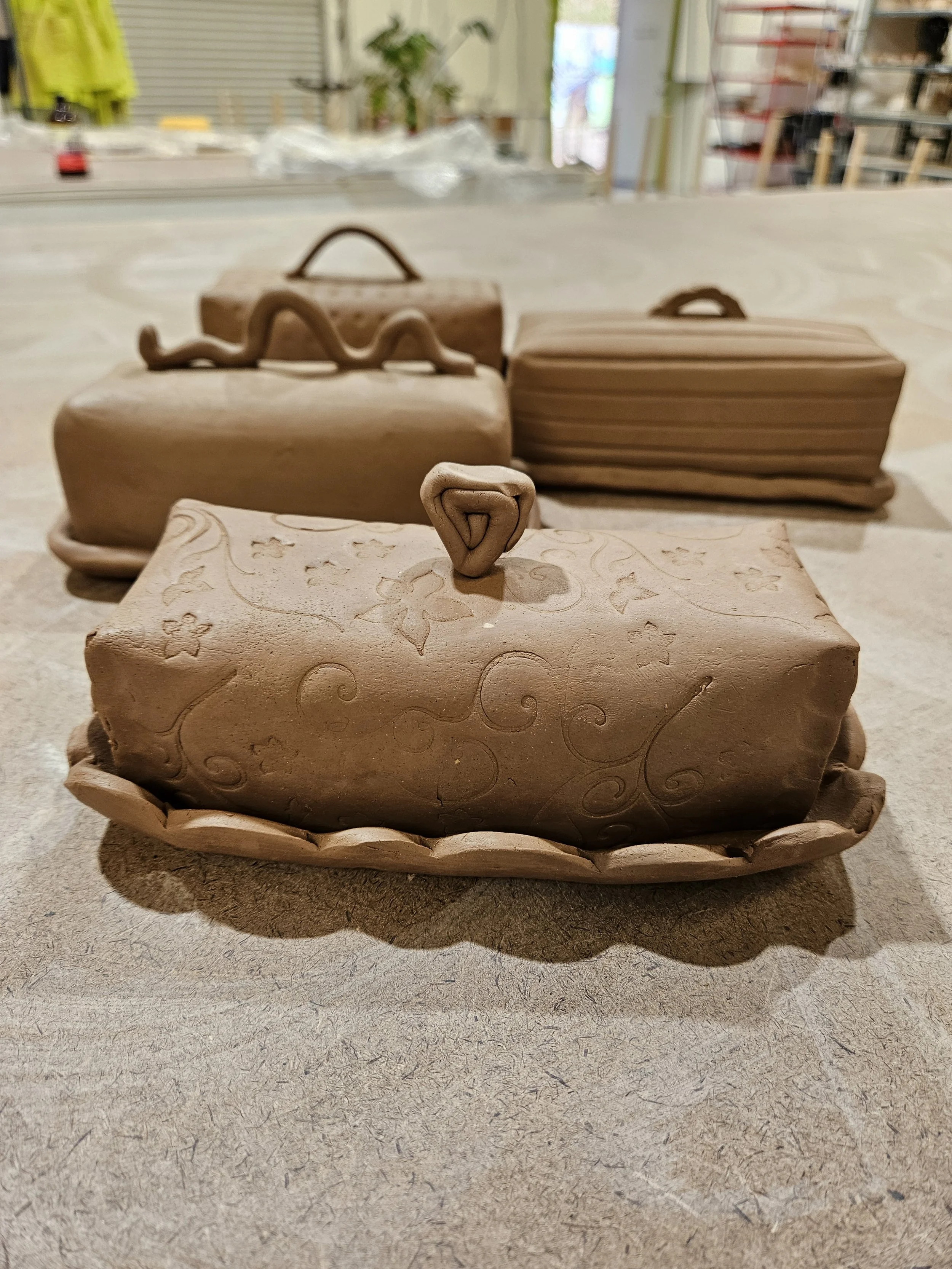 Four handcrafted clay butter dishes, each with different decorative patterns, placed on a wooden table in a studio setting.