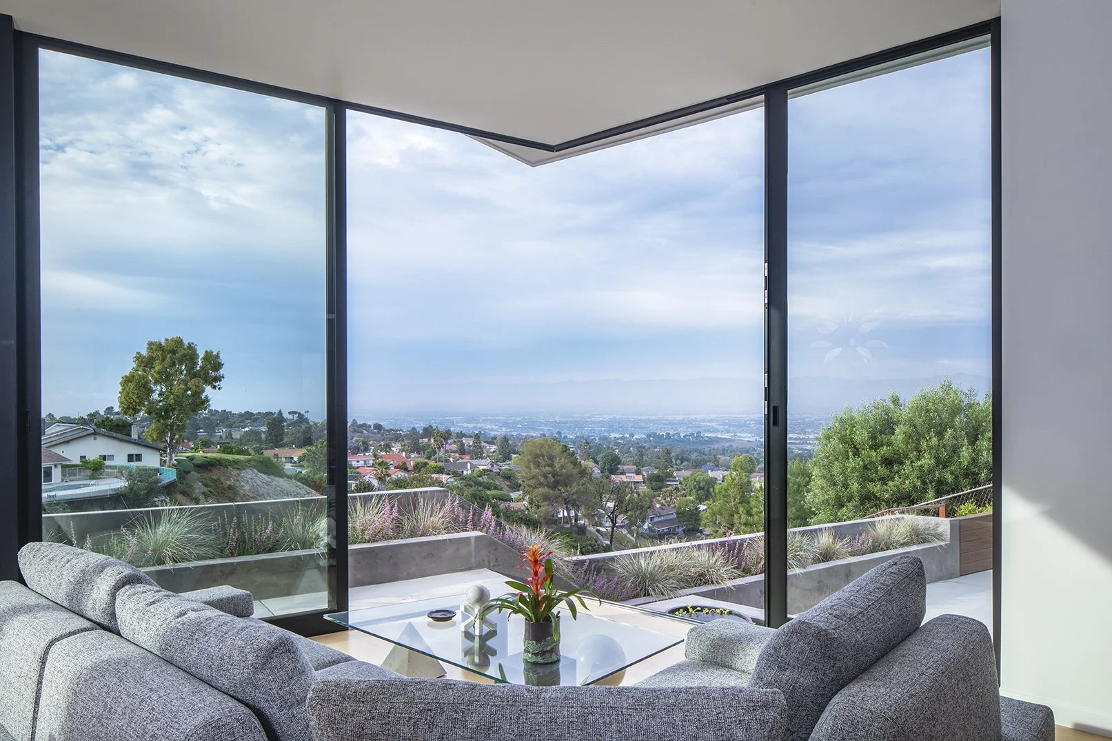 Living room with large floor-to-ceiling windows offering a view of los angeles landscape and distant mountains, with a gray sofa, a glass coffee table with a potted plant, and other furniture.