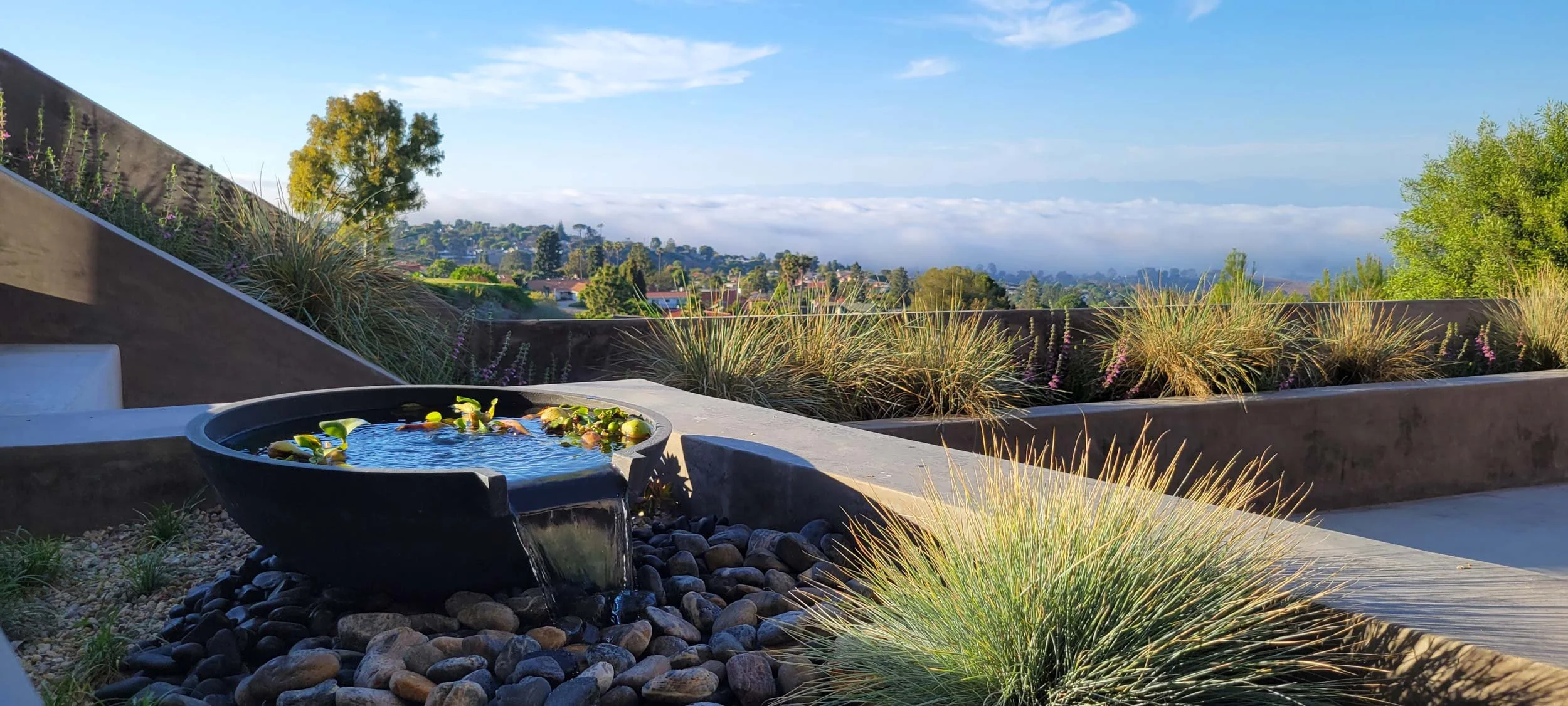 A modern outdoor patio with a water fountain, surrounded by decorative rocks and drought-tolerant plants, overlooking a suburban neighborhood with rolling hills in the background.