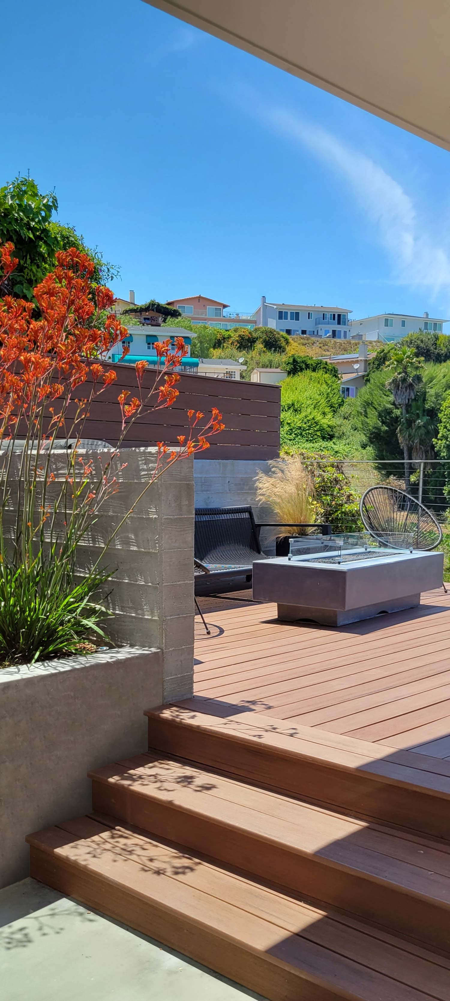 Outdoor patio with wooden stairs, plants, black chairs, a glass table, and a view of houses on a hillside under a blue sky.