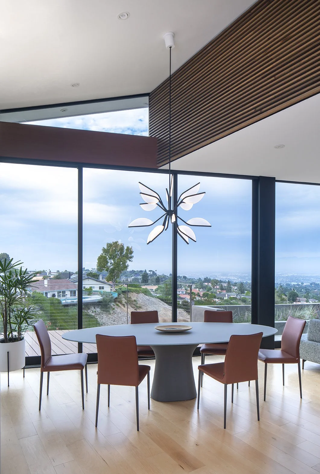 Modern dining room with a round table surrounded by six brown chairs, large floor-to-ceiling windows with a scenic view, a hanging ceiling light resembling a flower, and a potted plant in the corner.
