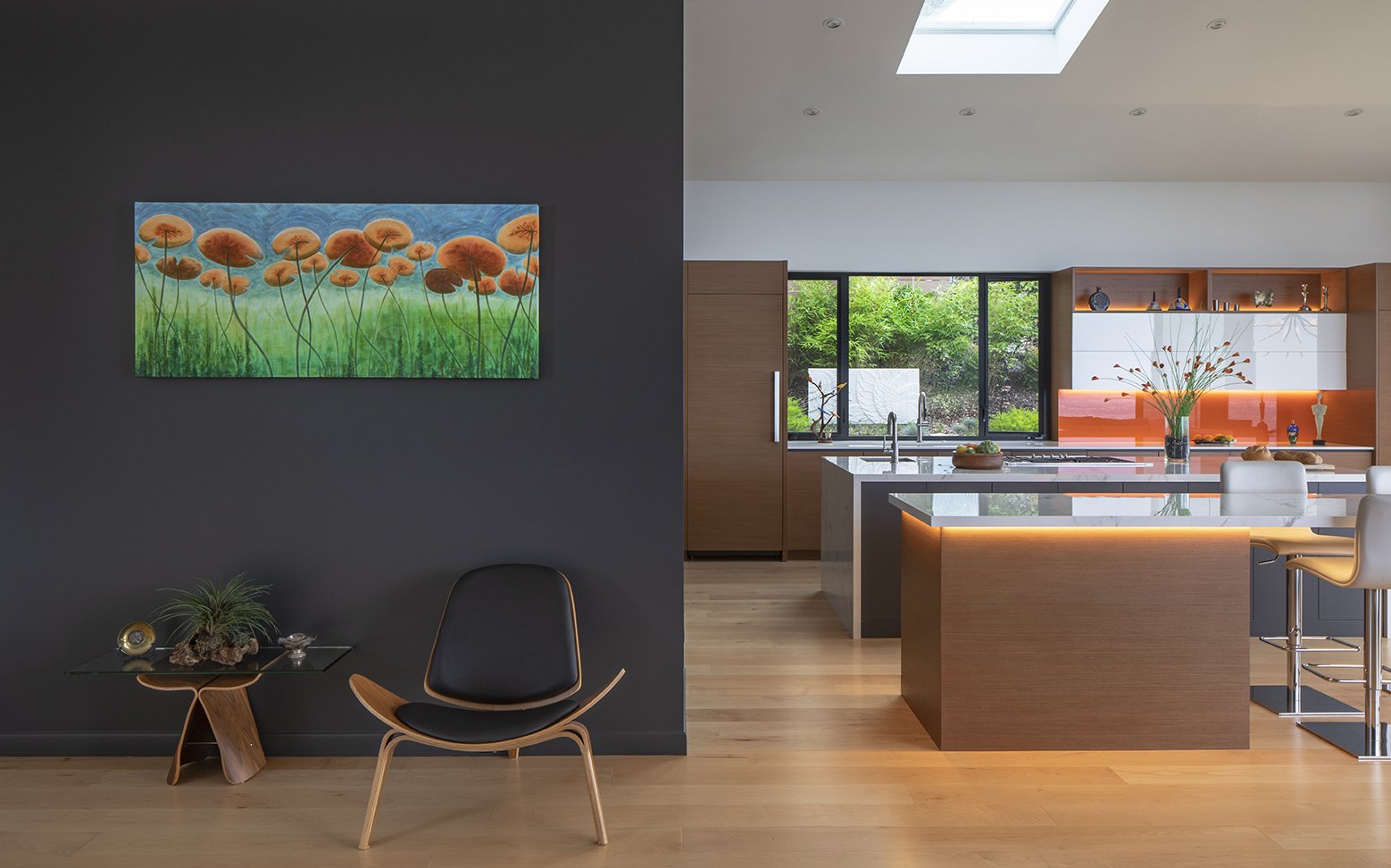 Modern kitchen and dining area with wooden cabinets, white chairs, a large island, and a window overlooking greenery; living space with a black wall, a colorful flower painting, and a small table with decorative items and a black chair.