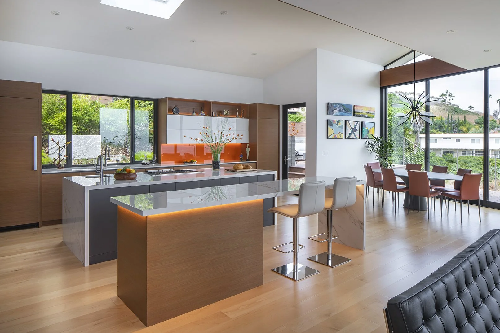 Modern open-concept kitchen with island, led strip lighting, white marble countertops, and dining area with large windows showing greenery and neighboring houses.