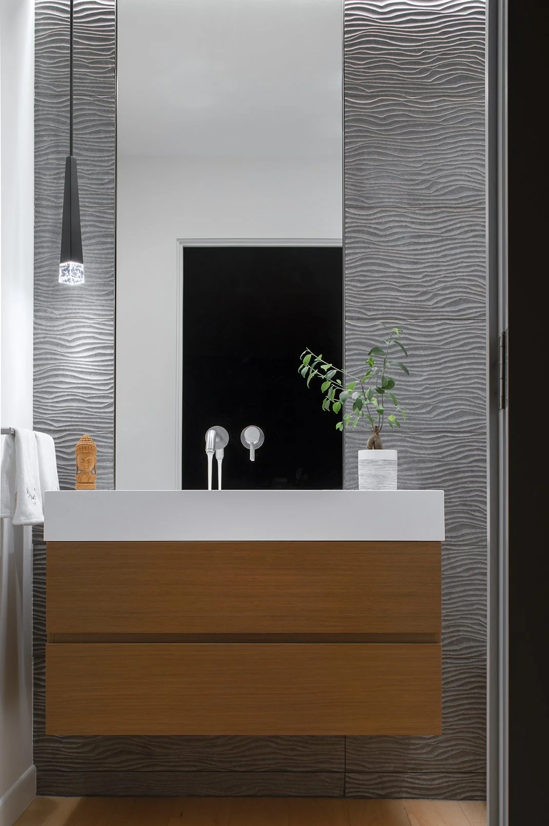 Modern bathroom floating vanity with a white sink, wooden drawers, brushed nickel faucet, a small potted plant, and a Buddha figurine, set against textured concrete wall tiles and a mirror in a powder room.