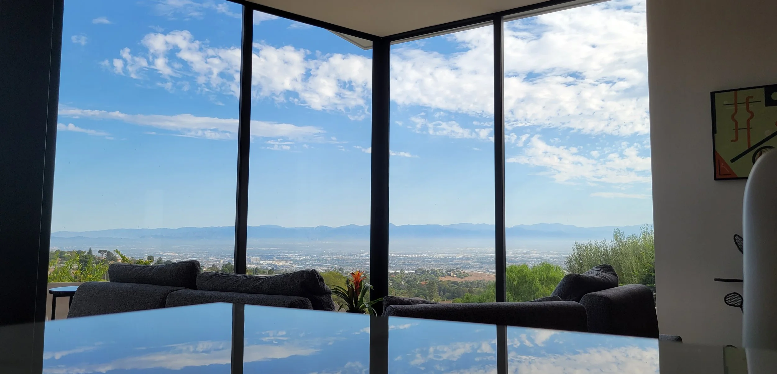 A modern interior room with large floor-to-ceiling windows overlooking a scenic landscape with mountains, sky, and clouds. There are chairs and a table inside, and a colorful abstract painting on the wall.
