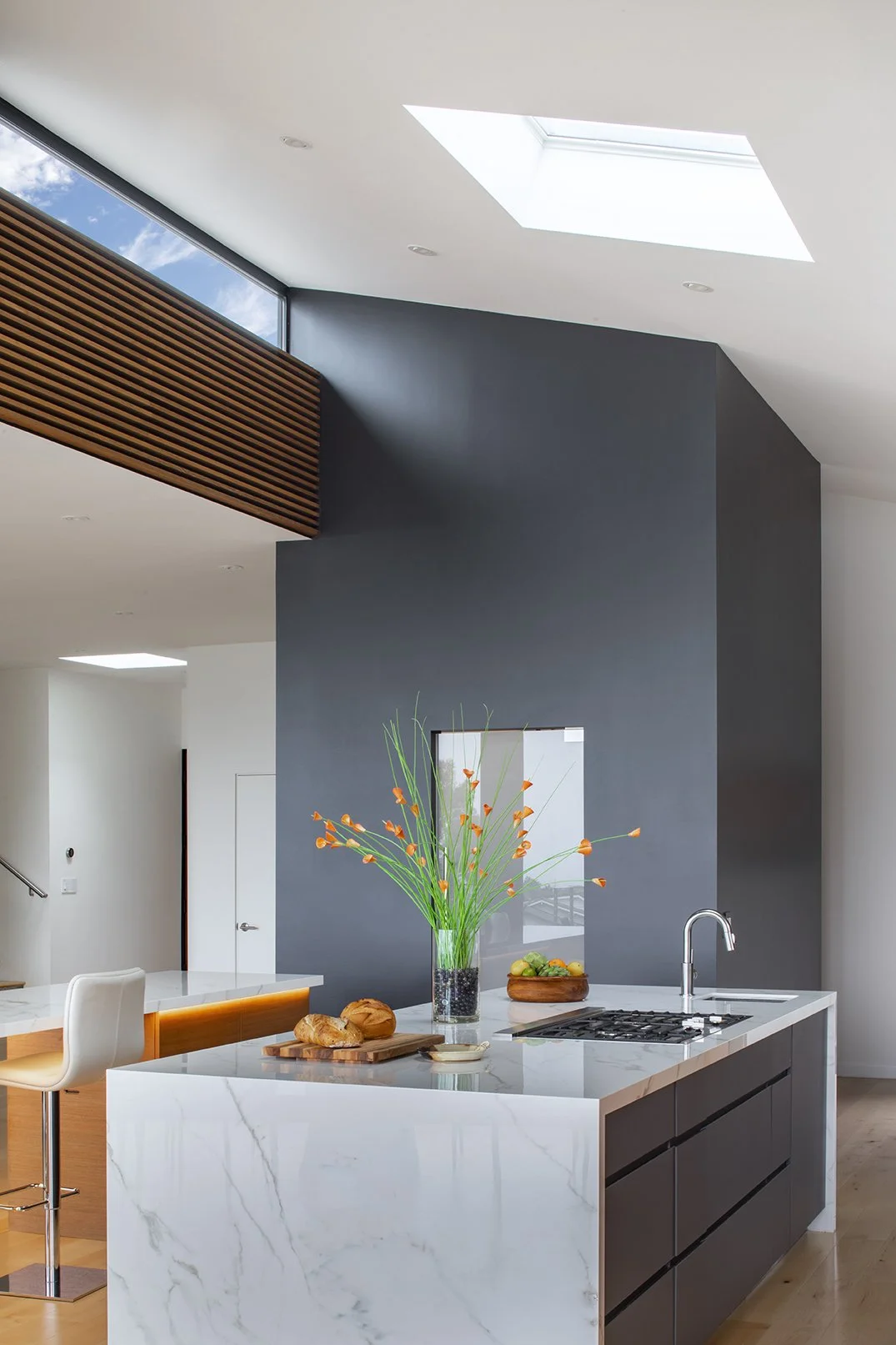 Modern kitchen with white marble island, black cabinets, gray accent pantry wall, vase with orange flowers, and skylights.