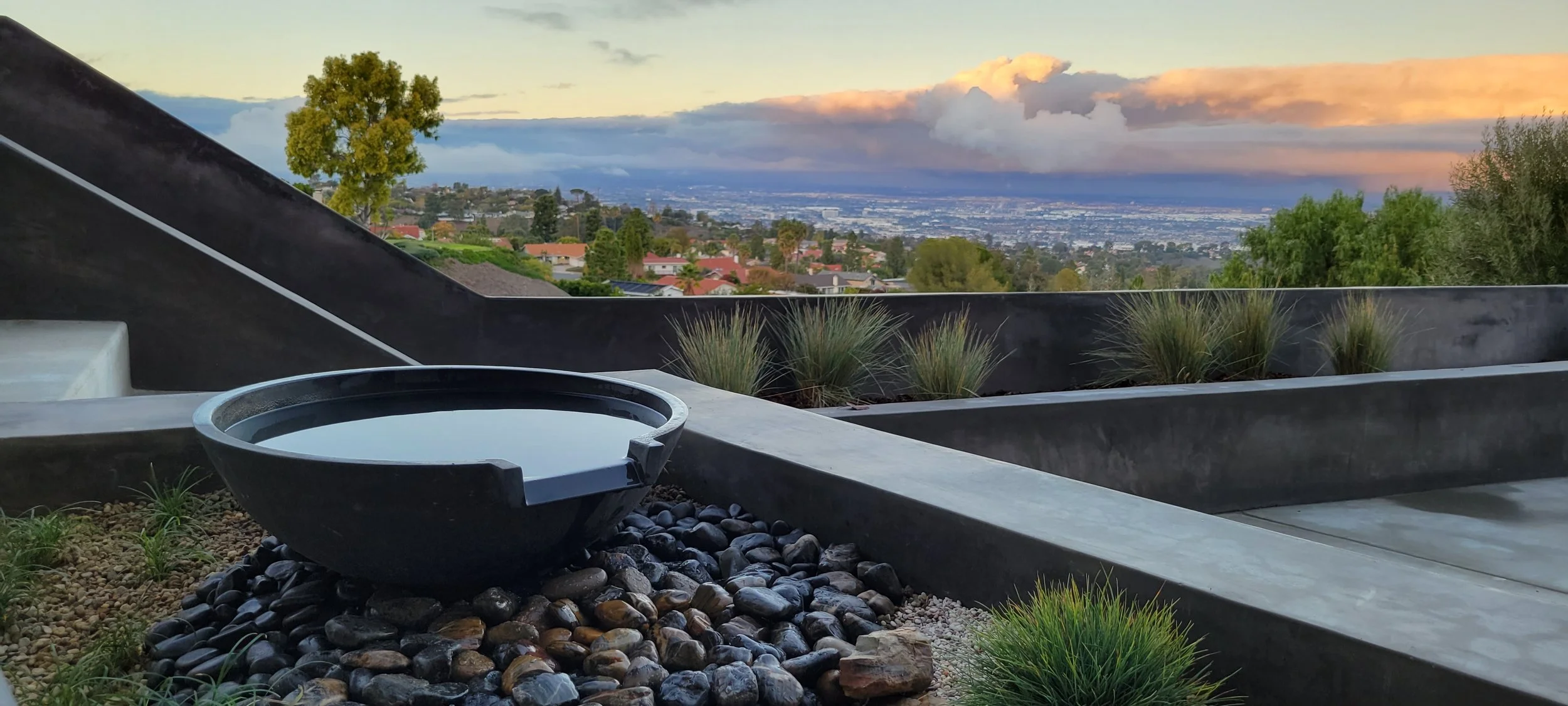 A modern outdoor terrace with a black bowl filled with water, black pebbles, and small green plants, overlooking a suburban landscape and a cloudy sky at sunset.