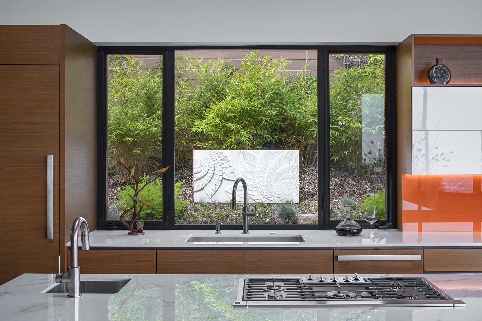 Modern kitchen with a large window looking onto bamboo, carved marble sculpture, countertop with a sink, decorative items, and a island stove.