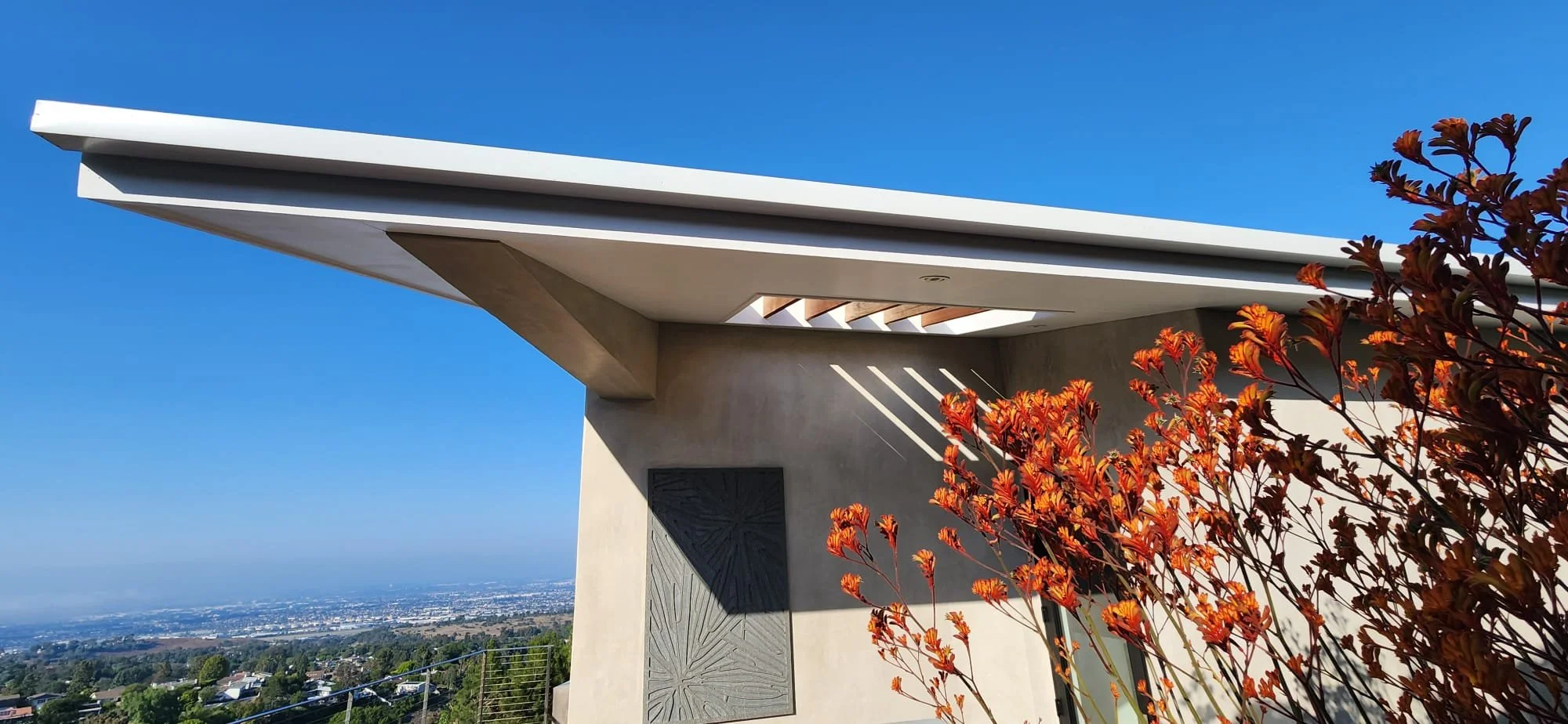 Modern house with an overhanging trellis roof, decorative carved wall panel, kangaroo paws, overlooking a cityscape under a clear blue sky in los angeles.