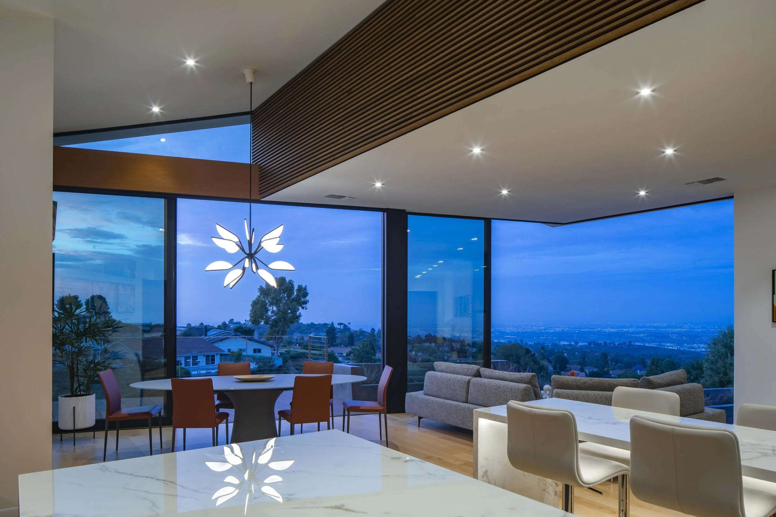 Modern living room with floor-to-ceiling windows showing a twilight sky and neighborhood view. Features a dining table with orange chairs, a gray sectional sofa, and a white armchair. A decorative light fixture resembling a butterfly is hanging from 