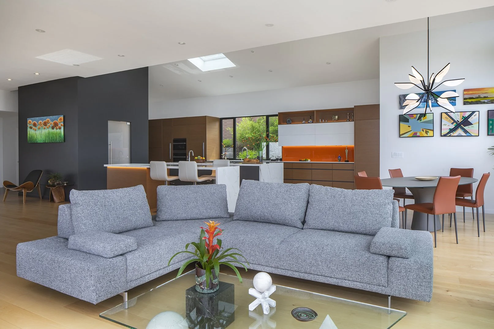 Modern open-concept living room and kitchen with gray sofa, colorful paintings, orange chairs around a dining table, and a black coffee table with decorative items.