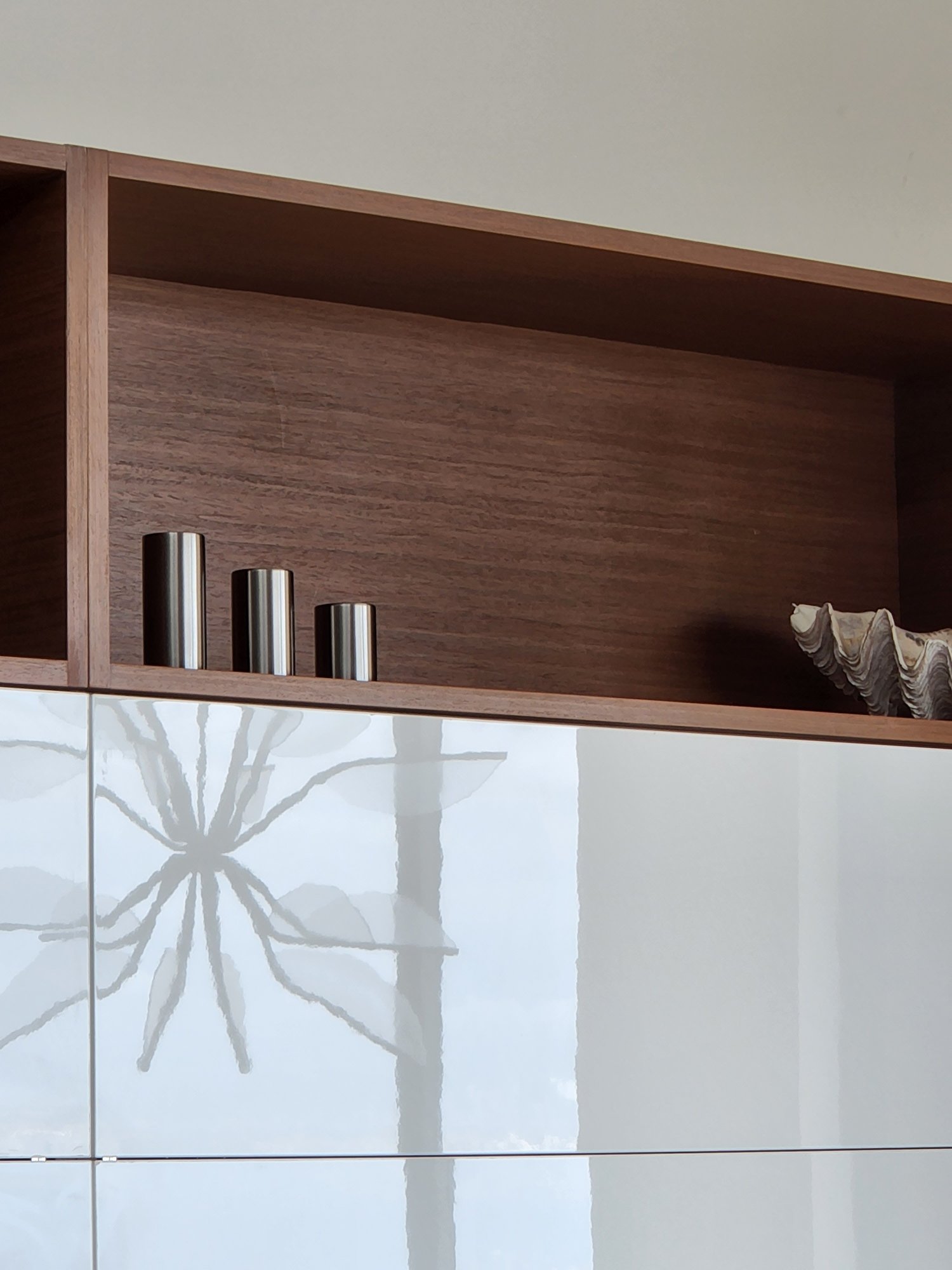 Wooden kitchen cabinet shelf with three metal canisters and a pile of napkins reflected on a shiny white surface.