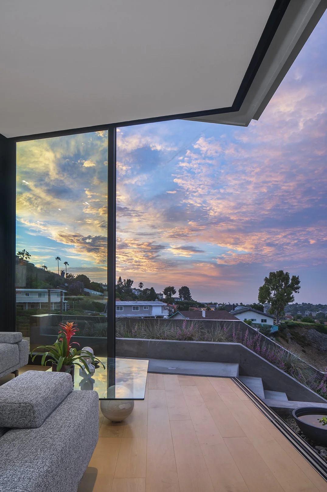 Interior view of a modern living room with wooden floor and large glass windows showing a spectacular sunset sky with clouds and trees outside.