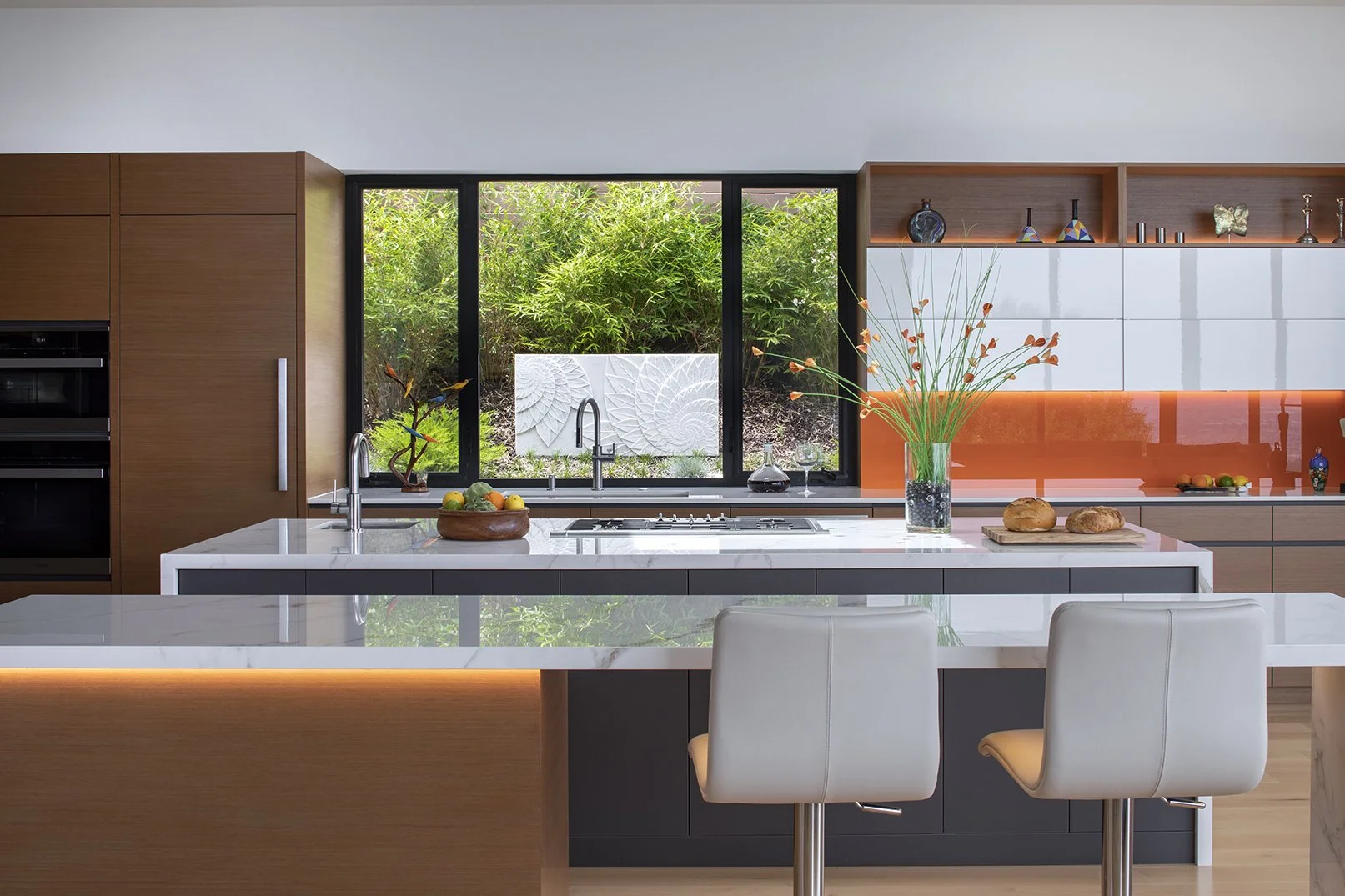 Modern kitchen with white marble island, wooden cabinets, large window with greenery, decorative vases, and fresh bread on a tray.