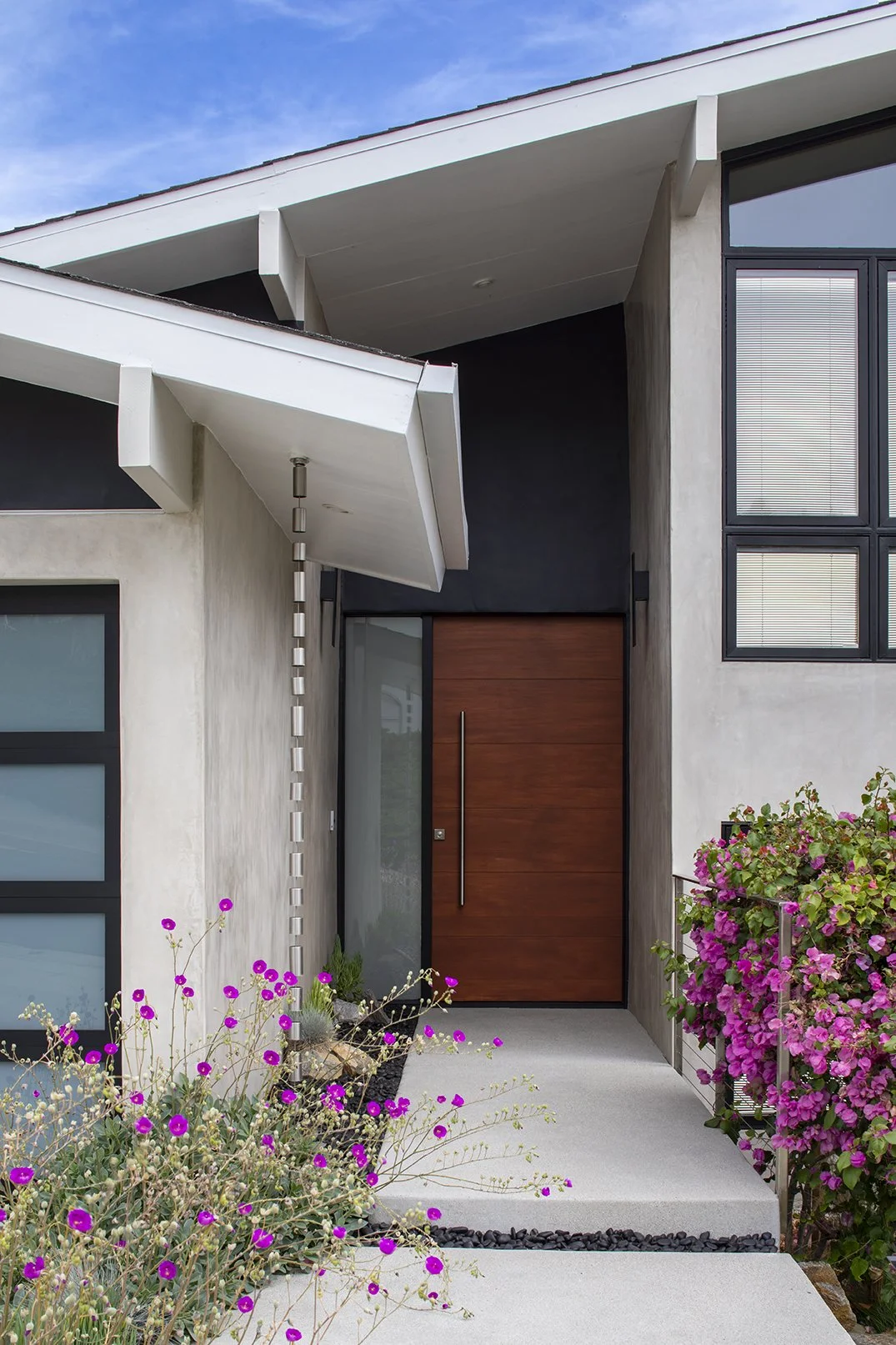Modern house entrance with wooden door, large glass window, rain chain, glass garage door and colorful flowers in the garden.