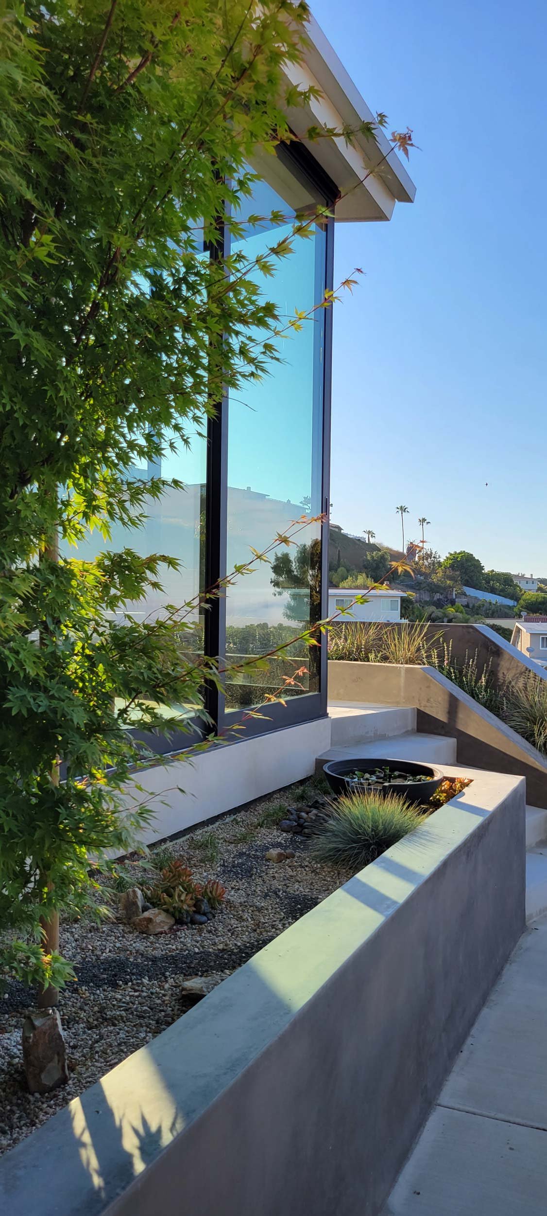 Modern house with large glass windows, a landscaped terrace with plants, and a hillside in the background under a clear blue sky.