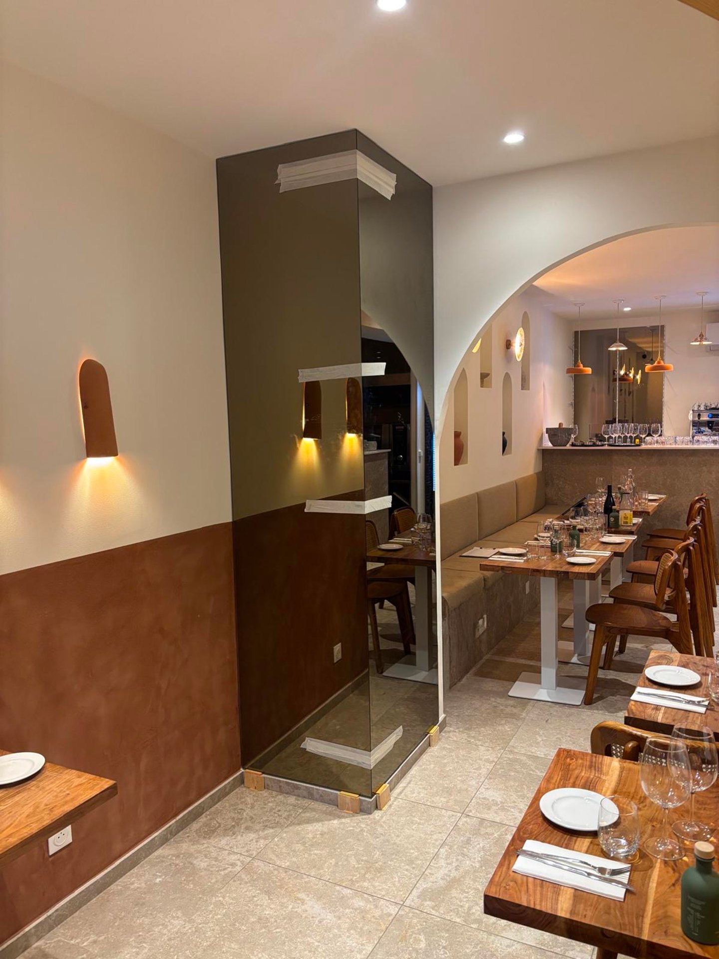 Intérieur d'un restaurant avec des tables en bois, des verres, assiettes, et un mur avec des niches décoratives, éclairages suspendus, et une zone bar en arrière-plan.