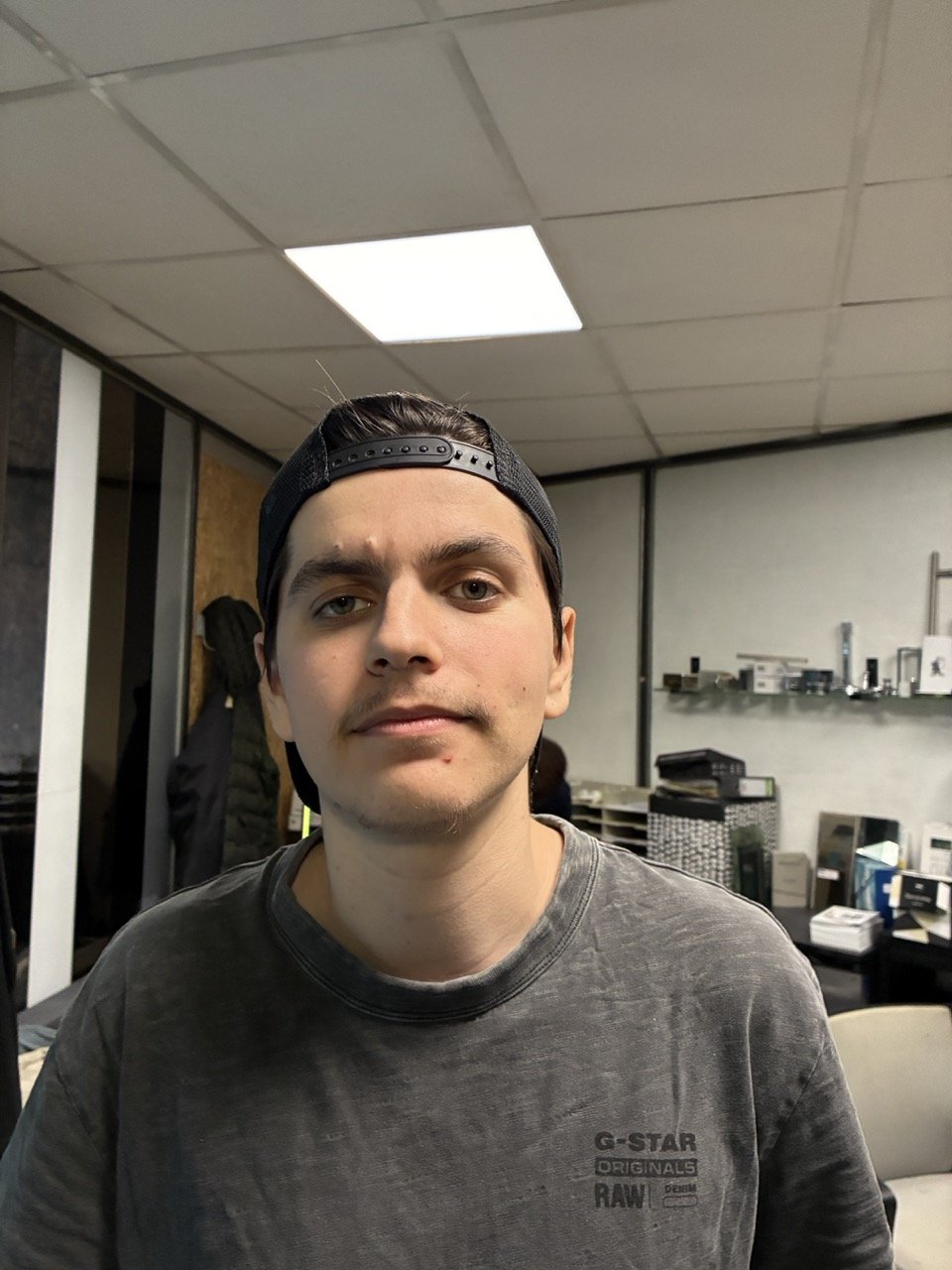 Jeune homme portant une casquette noire, en haut d'une tête, souriant légèrement, dans une pièce avec un plafond lumineux et divers objets sur une étagère en arrière-plan.