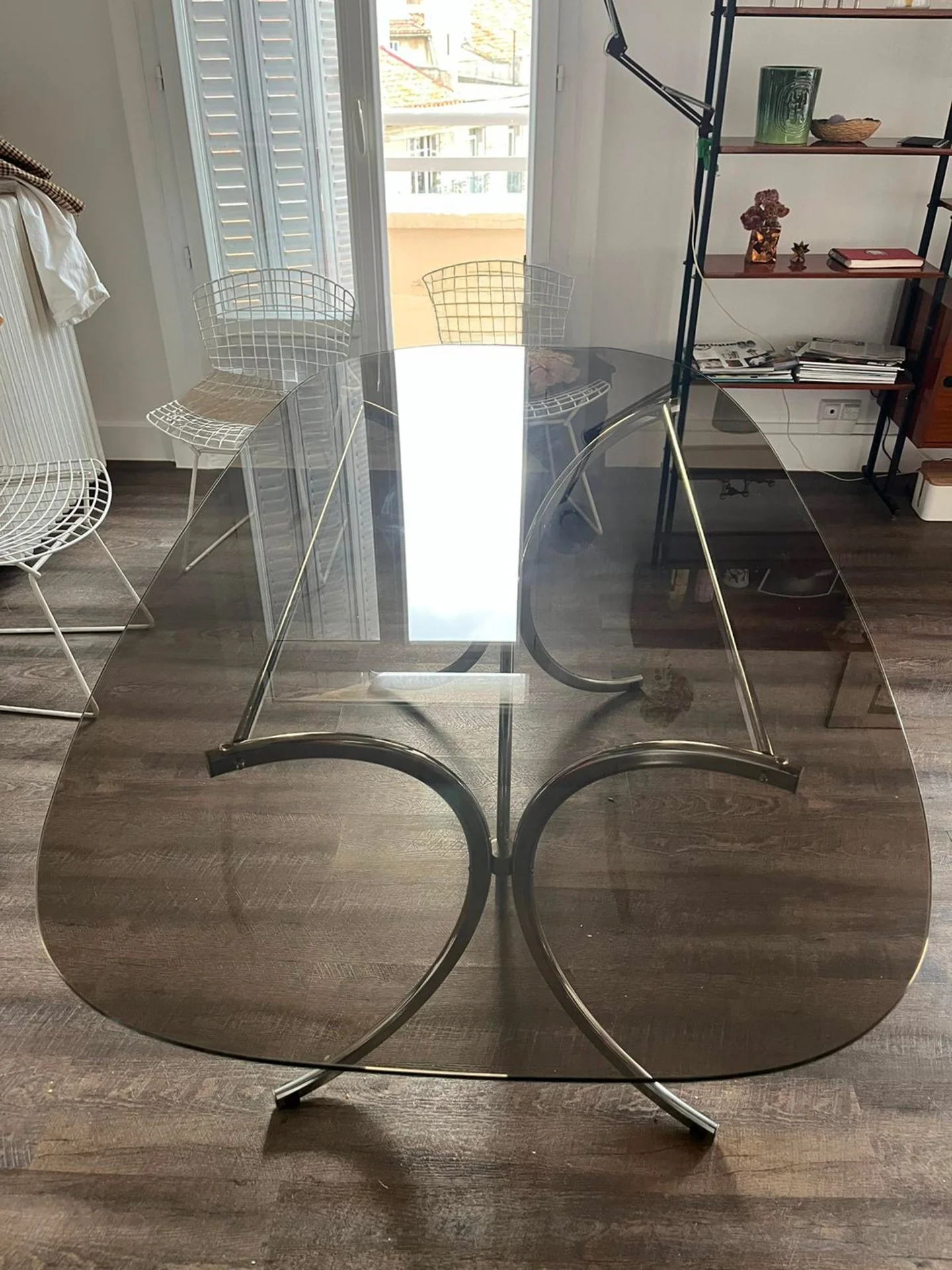 Plateau de table en verre avec structure en métal noir, placé dans une pièce avec parquet en bois, fenêtre avec volets battants et étagère en métal et bois avec diverses décorations et livres à Marseille