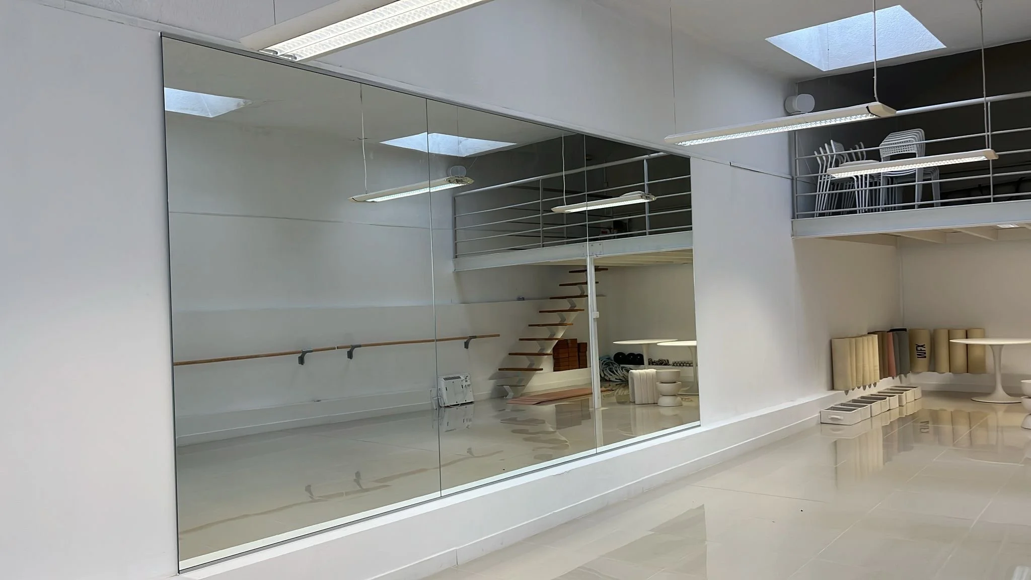 Intérieur d'une salle de sport avec un grand miroir, des étagères pour tapis de yoga, un mur blanc avec des chaises empilées, et des équipements sportifs comme des sacs de frappe.