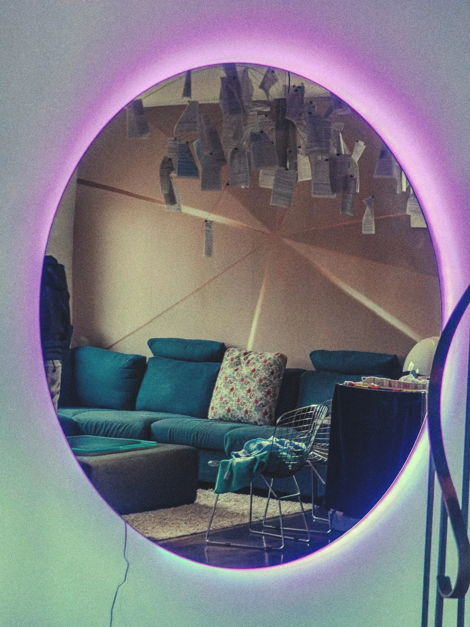 Un miroir rond avec une lumière rose et verte autour, reflétant un salon avec un canapé bleu, un coussin fleuri, une table basse, une chaise en métal, un tapis beige, et un mur décoré de papiers suspendus.