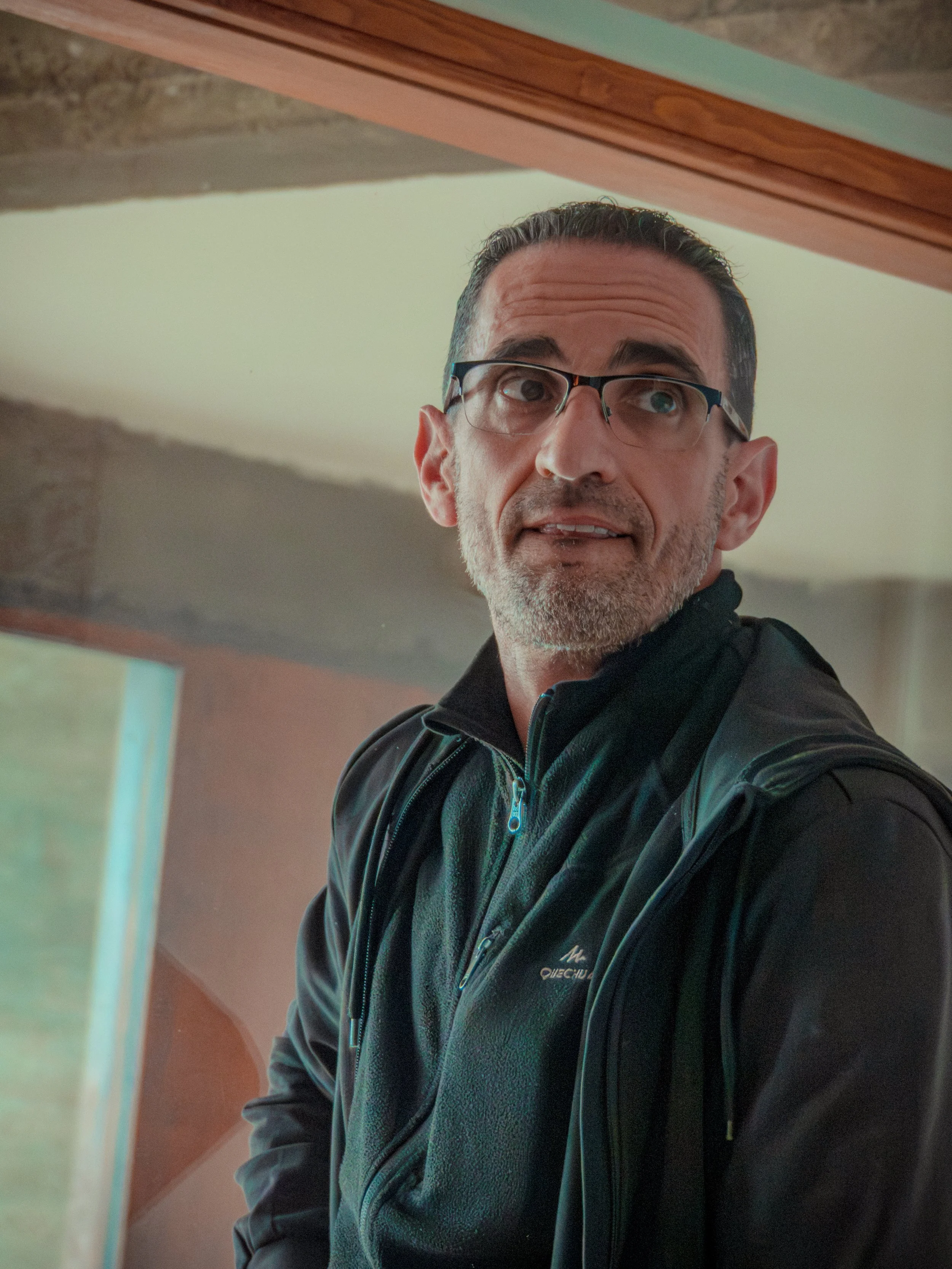 Charlot portant des lunettes et une veste noire, vu dans un miroir, avec une surface en bois et un mur beige en arrière-plan.