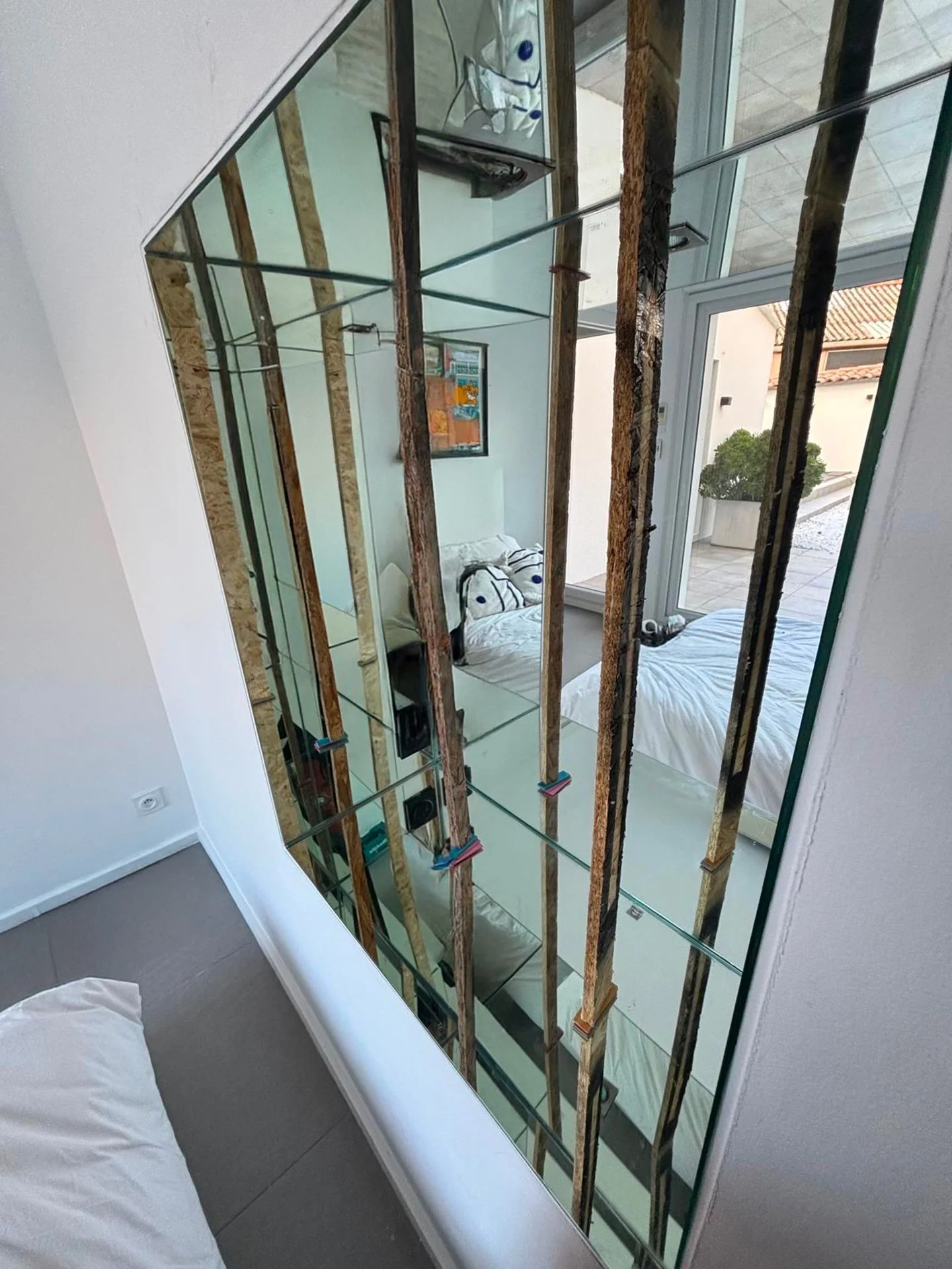 Un miroir ancien en cours de restauration, avec des échelles en bois et des outils visibles, reflétant une pièce avec un lit et des fenêtres donnant sur une terrasse extérieure.