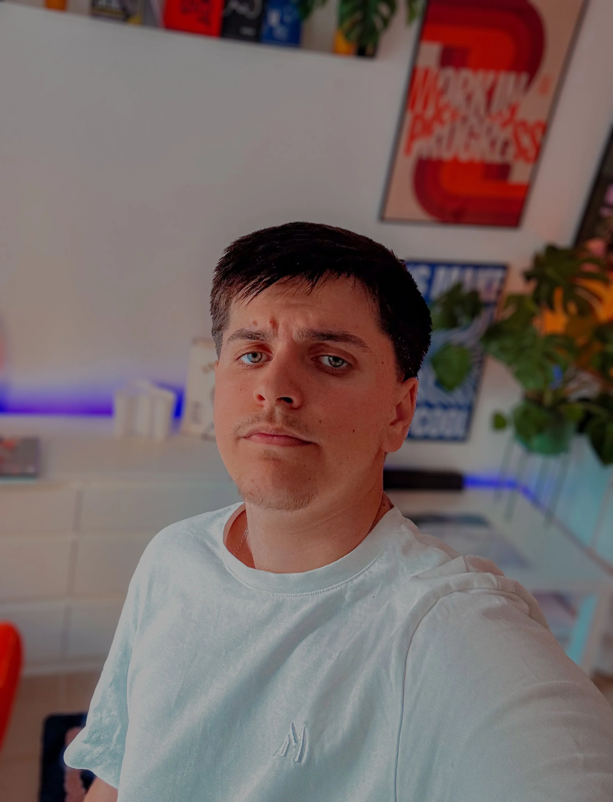 Guy à la peau claire et aux yeux bleus portant un t-shirt blanc, avec un mur décoré de posters colorés derrière lui.