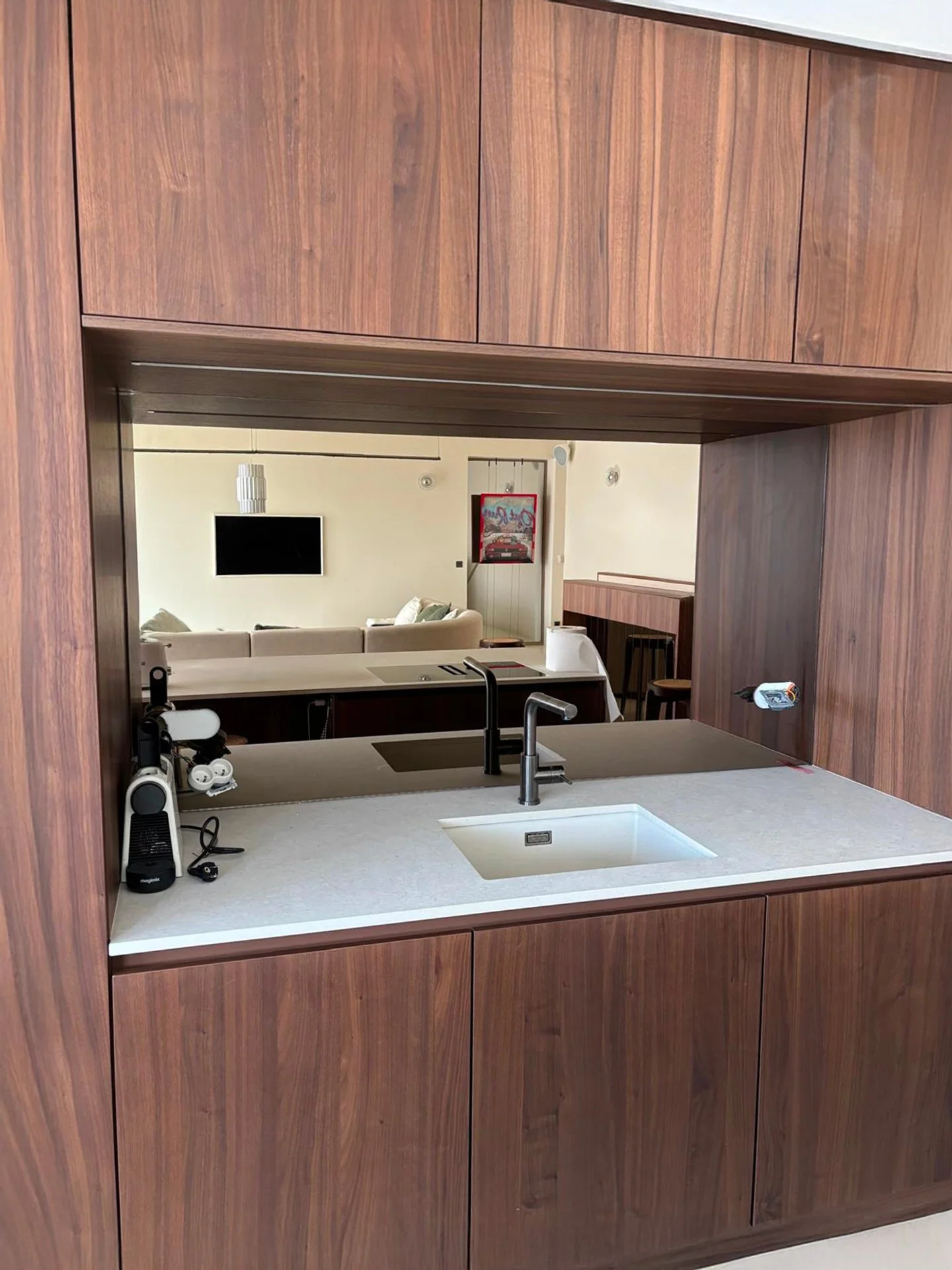 Vue de face d'une cuisine avec un comptoir blanc, un évier, un robinet noir, des meubles en bois foncé, un plan de travail, un fond blanc avec une télévision et un tableau.