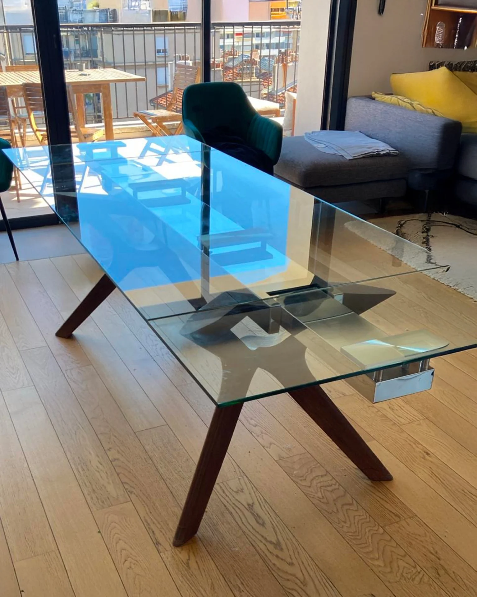 Une table en verre blanc avec pieds en bois foncé, située dans une pièce avec un canapé gris, des coussins jaunes, et un balcon avec une table en bois et des chaises, en arrière-plan.