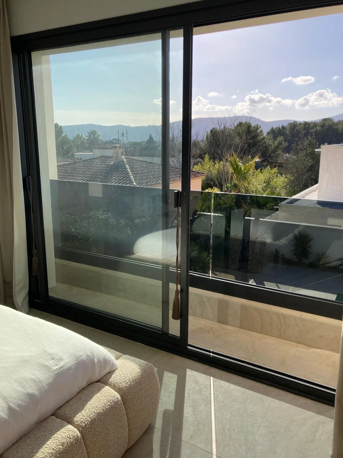 Une vue depuis une pièce intérieure avec une grande fenêtre en verre coulissante, montrant un balcon avec des plantes et un paysage de montagnes et de maisons sous un ciel ensoleillé.