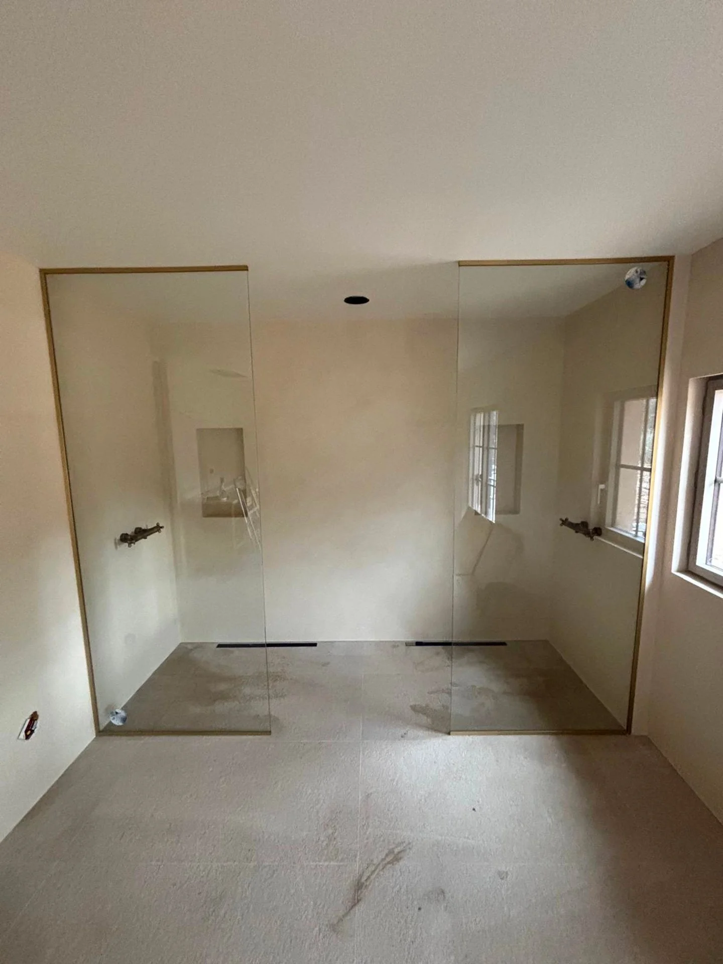 Une pièce en cours de travaux avec un mur blanc, deux fenêtres, un sol en béton et une cloison en verre.