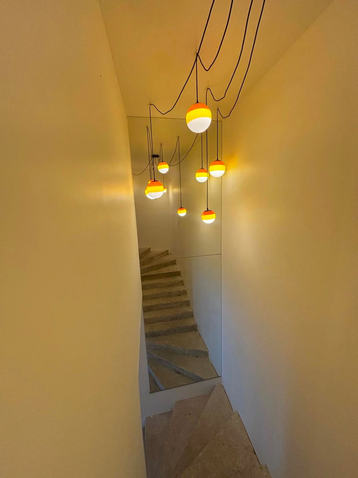 Un escalier en marbre, vu depuis un couloir avec des murs jaunes, éclairé par des lampes suspendues à des fils noirs zigzaguant au plafond.