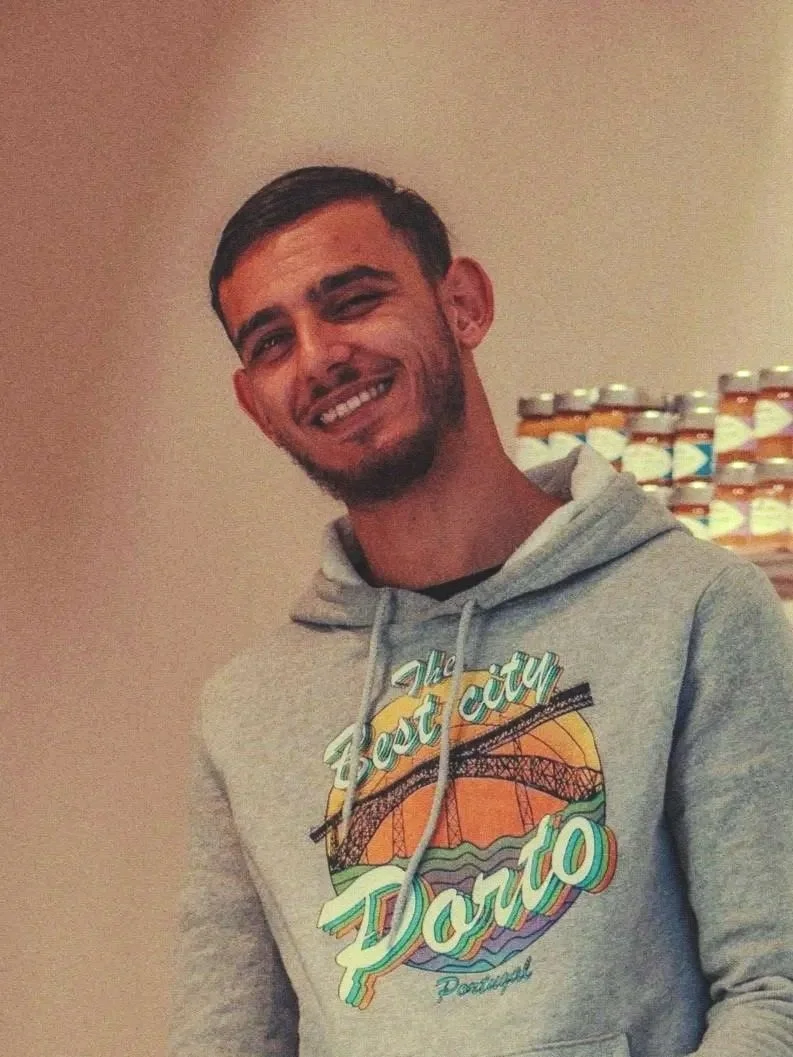 Jeune homme souriant portant un sweat à capuche gris avec un design de pont et coucher de soleil, dans un intérieur