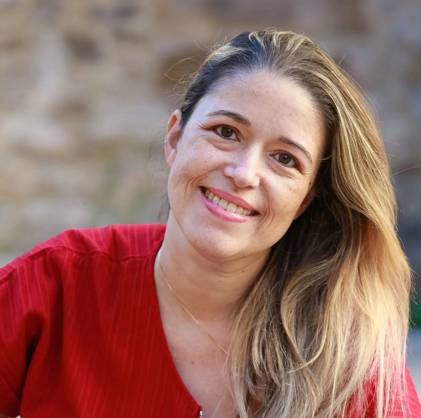 Femme souriante portant un haut rouge, avec des cheveux longs, bruns et blonds, en arrière-plan flou.