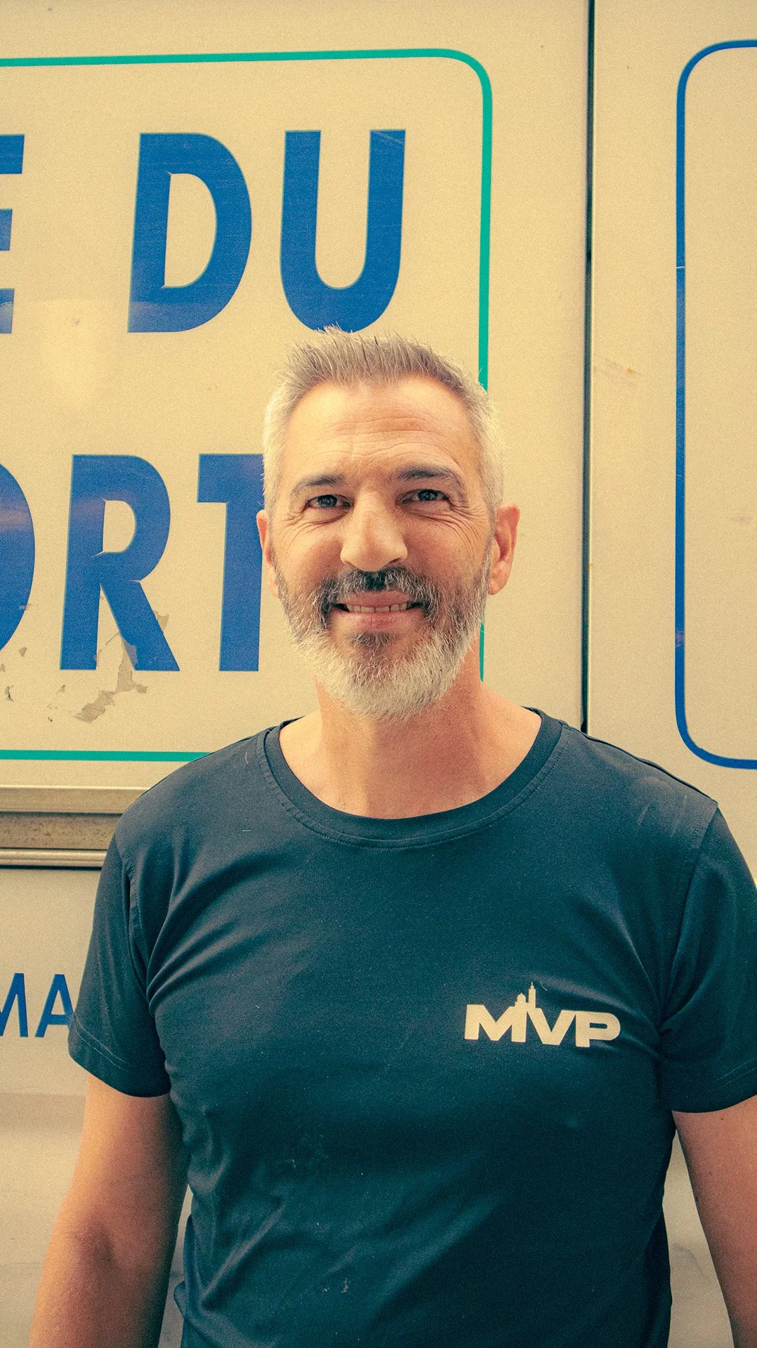 Un homme souriant avec une barbe et des cheveux gris, portant un t-shirt noir avec le logo MVP, devant une affiche bleue avec des lettres françaises.