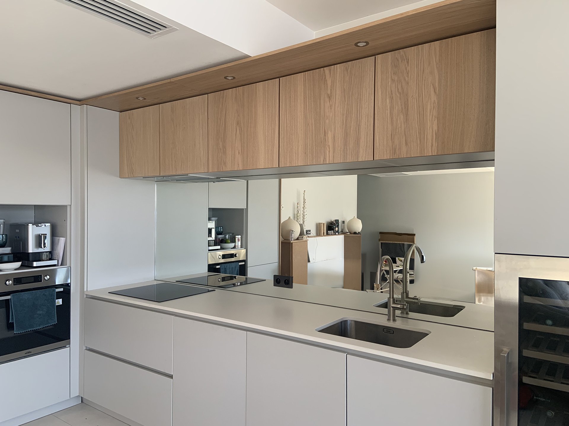 Cuisine moderne avec îlot blanc, miroir argenté clair 6mm avec bords polis et trous pour prise, évier, cocotte-minute, four, armoires en bois clair, décorations minimalistes, et un coin salle à manger visible au fond.