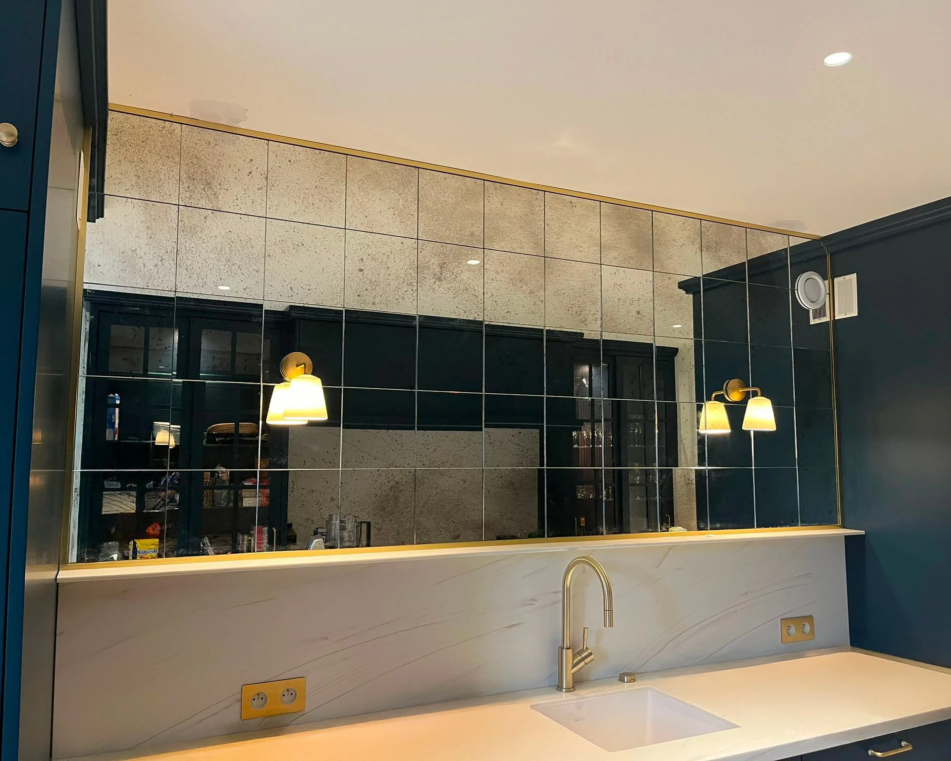 Un miroir mural en carreaux noirs dans une cuisine, reflétant une porte vitrée et un mur en pierre. La surface du miroir est divisée en plusieurs sections, encadrées par des joints en métal. Il y a des lampes suspendues avec abat-jour jaunes, un évie