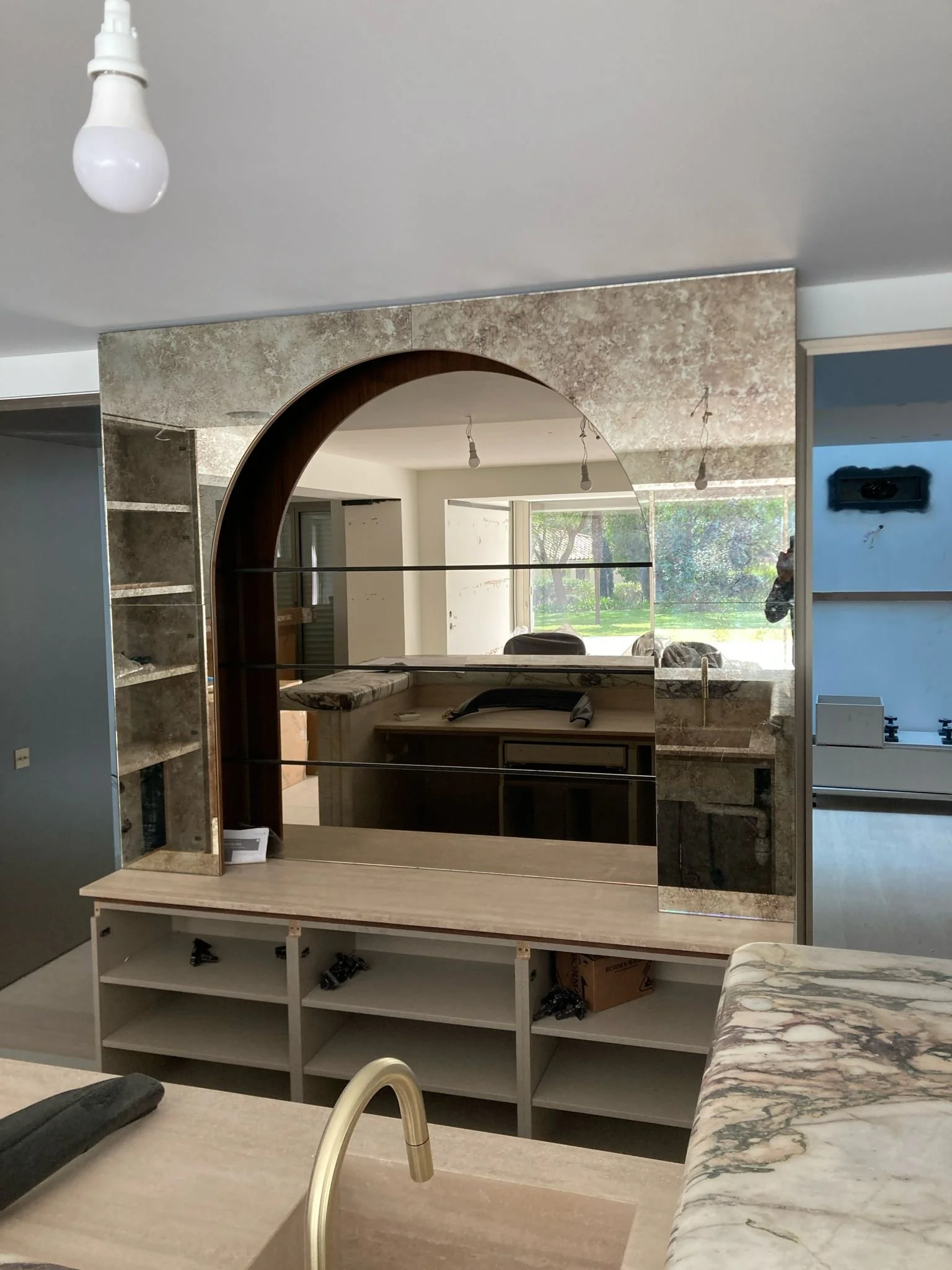 Une grande pièce en cours de rénovation avec un miroir accroché au mur. La pièce a des fenêtres laissant entrer beaucoup de lumière et montre une vue de l'extérieur avec des arbres. Il y a un comptoir en marbre dans la pièce et des meubles en cours d'installation, avec des outils et des matériaux de construction visibles.