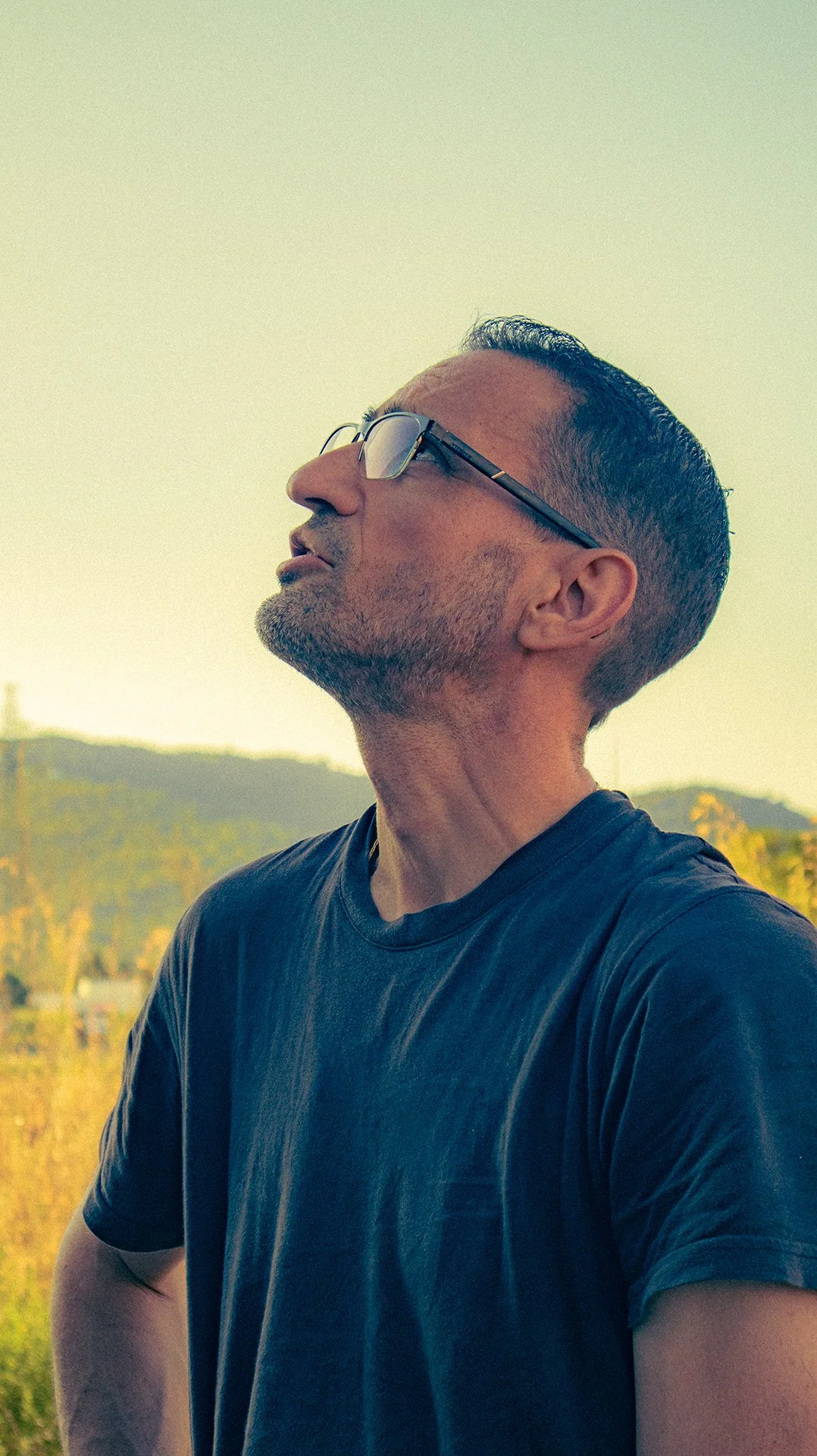 Un homme portant des lunettes, regardant vers le haut, en plein air avec un paysage de collines en arrière-plan.