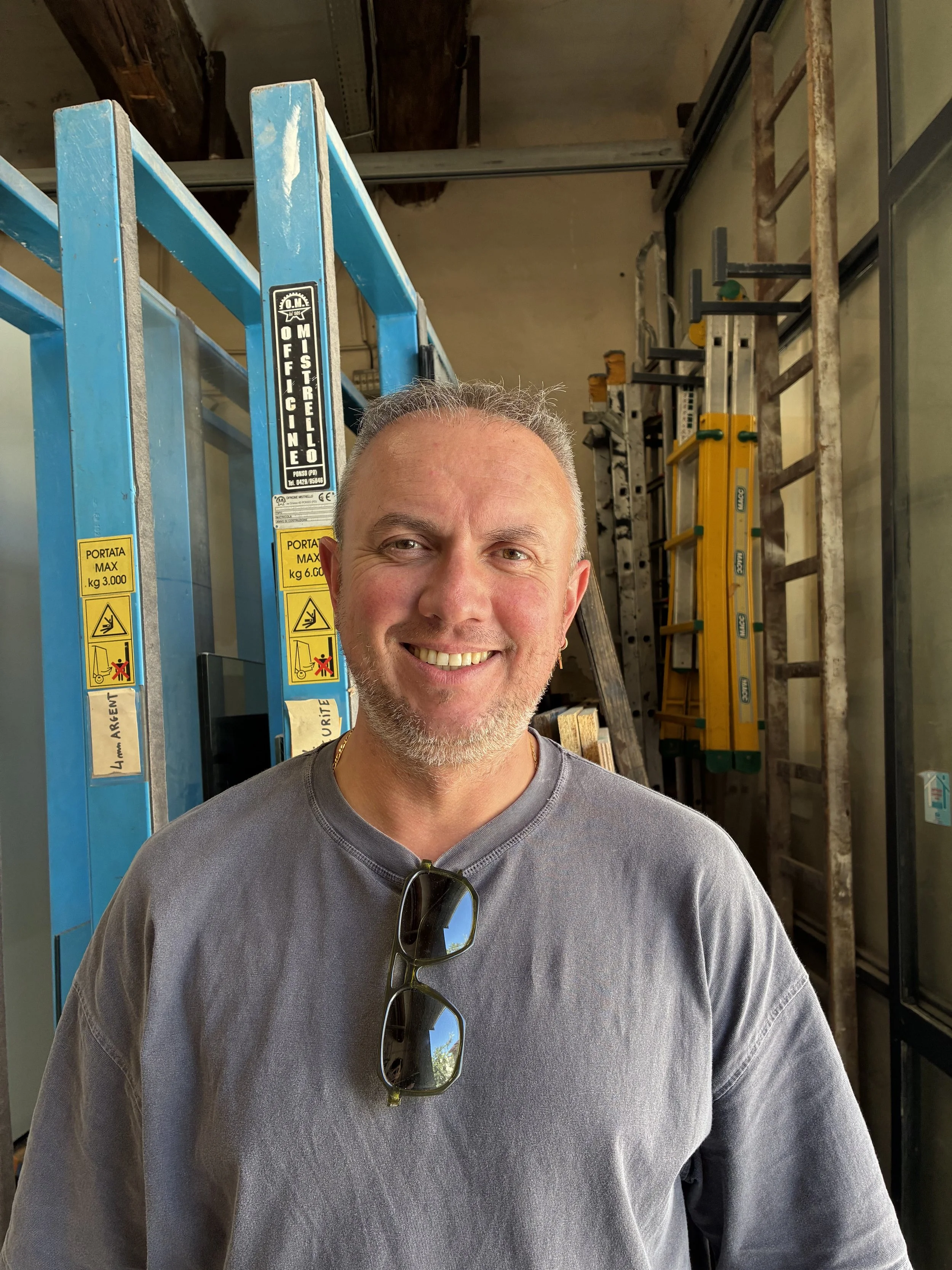 Un homme souriant portant un t-shirt gris, avec des lunettes suspendues à son cou, dans un entrepôt avec des échelles bleues et jaunes en arrière-plan.