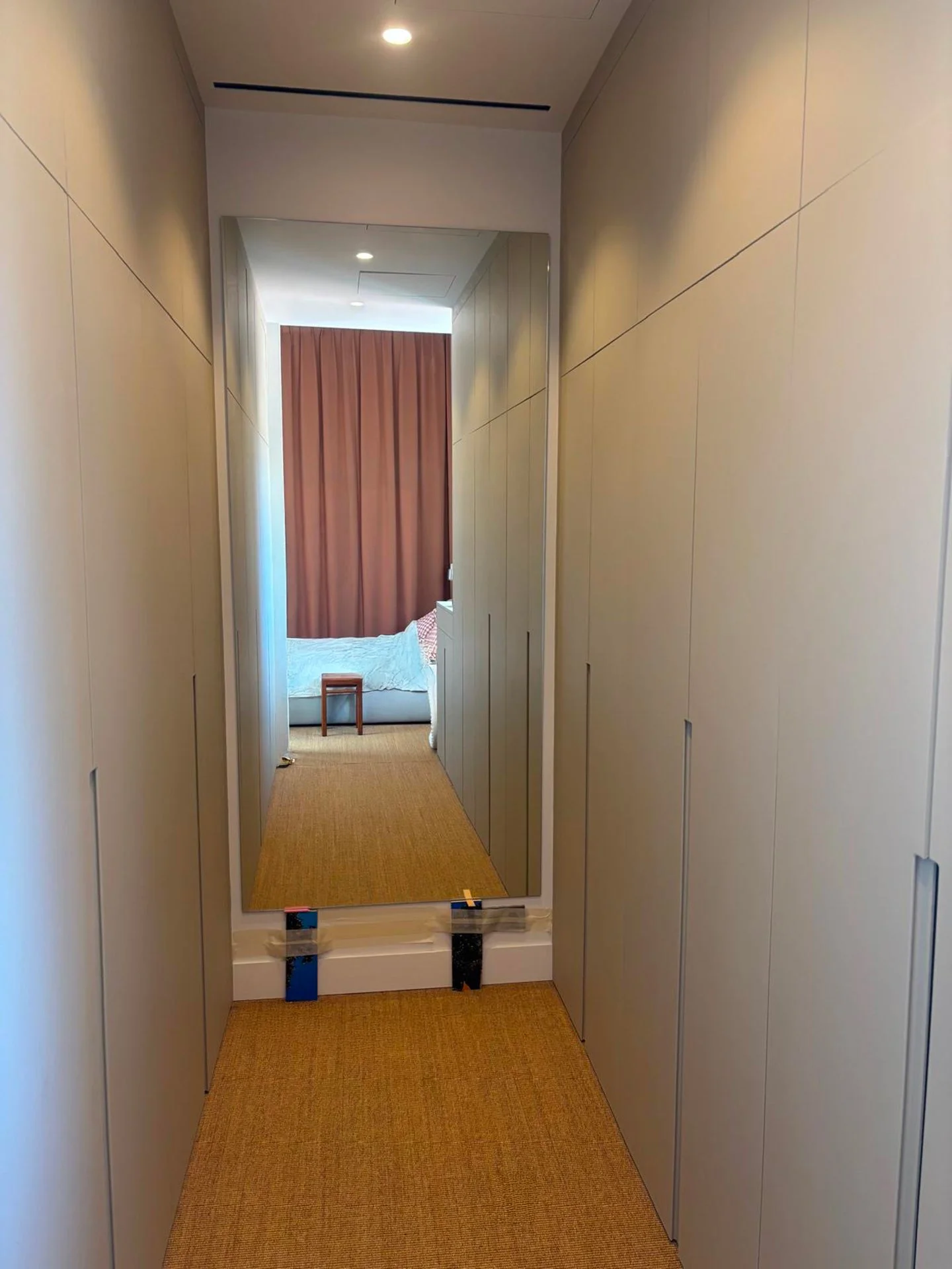 Un miroir dans un couloir d'hôtel, reflétant une chambre avec un lit, une table de chevet, un rideau marron et un tapis beige.