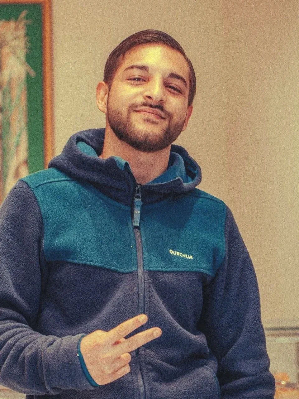 Jeune homme souriant portant une veste bleue foncée et un hoodie, faisant un signe de paix avec sa main, à l'intérieur.