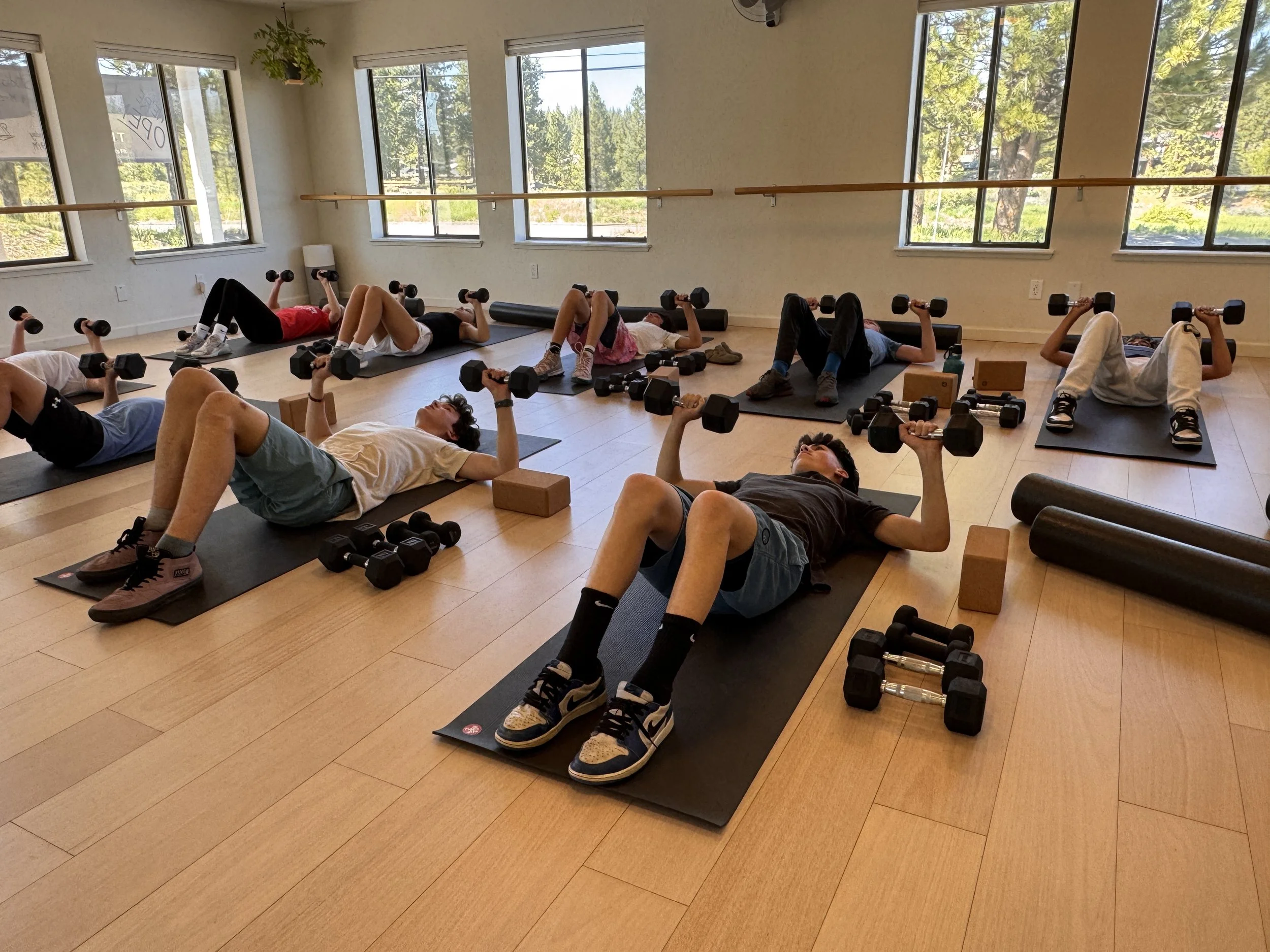 A group of children engaging in a workout class, lying on their backs on yoga mats, lifting dumbbells, and using foam rollers, with large windows revealing a green outdoor landscape.