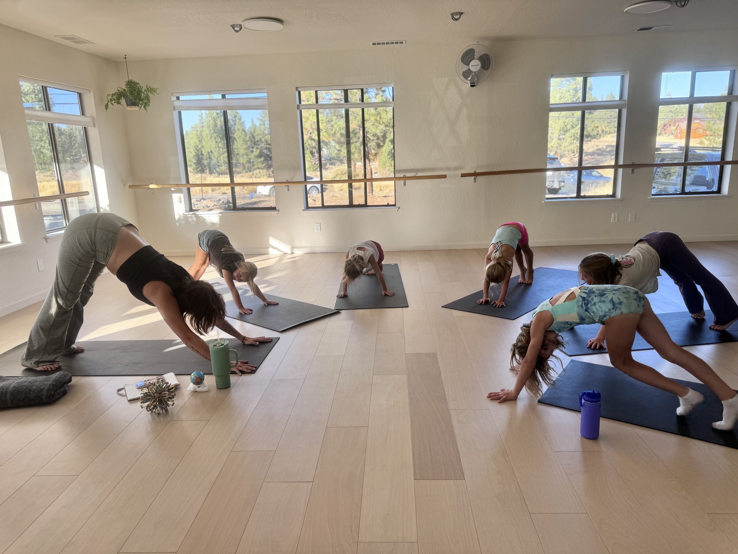 A yoga class with children practicing downward dog pose, led by an instructor in a bright studio with large windows, wooden floors, and a ballet barre.