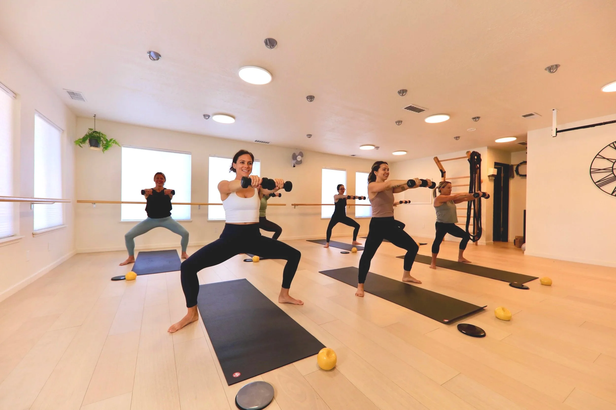 Group of women participating in a fitness class in a bright studio, doing squats with dumbbells on black exercise mats.